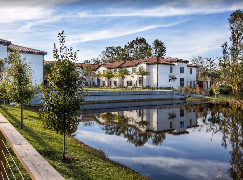 White two-story Mediterranean-style building with a red tile roof reflected in a pond, surrounded by trees and a grassy walkway.