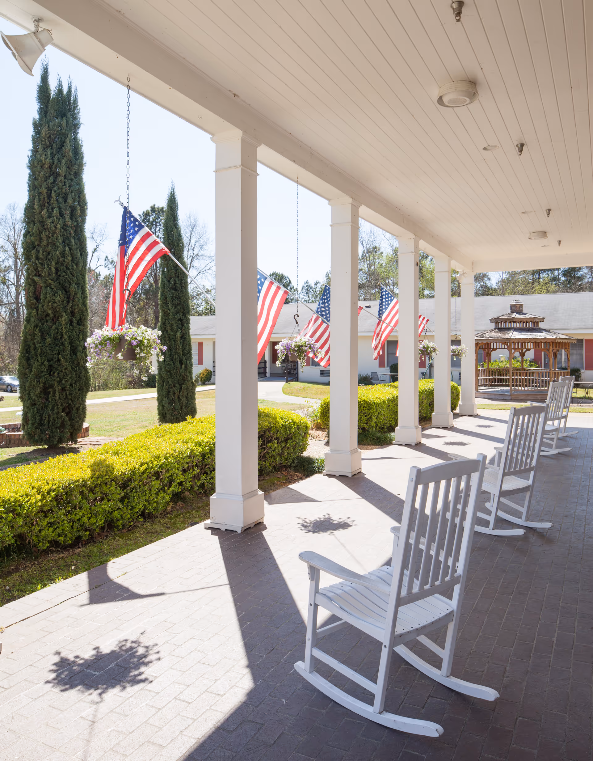 Covered outdoor walkway with white rocking chairs, American flags, and hanging flower baskets.
