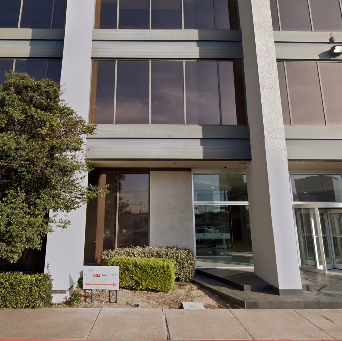 Modern office building entrance with glass doors and surrounding shrubbery.