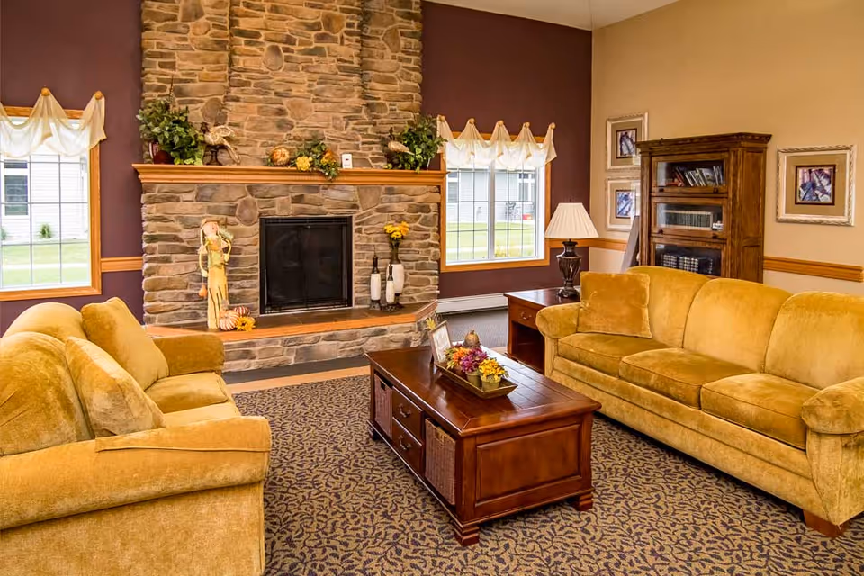 A cozy living room with two mustard yellow sofas facing each other, a wooden coffee table with a floral centerpiece, a stone fireplace decorated with plants and figurines, two windows with white curtains, a wooden bookshelf, and framed artwork on the walls.