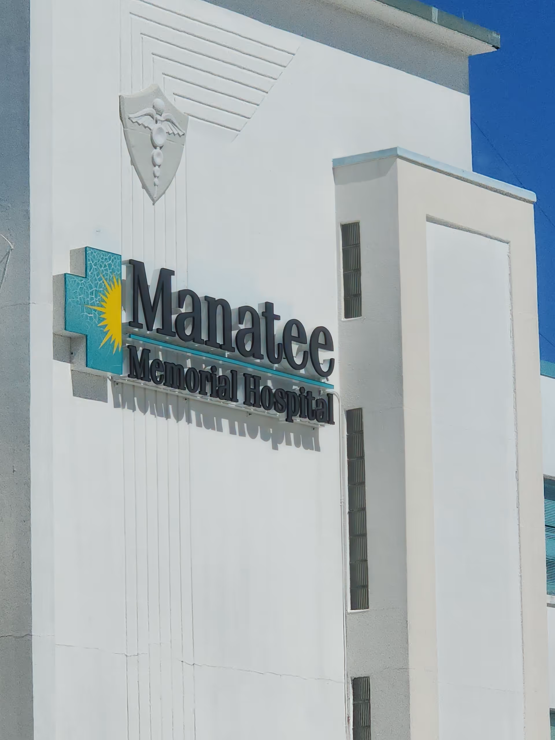 White hospital exterior with Manatee Memorial Hospital sign and crest on the wall.