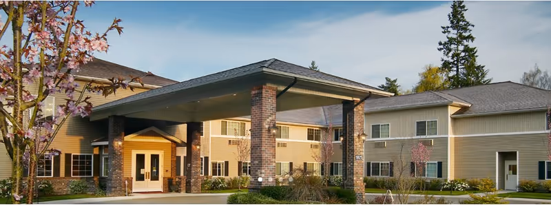 Exterior view of Normandy Park Senior Living entrance with a covered porte-cochere, brick columns, and landscaped grounds.