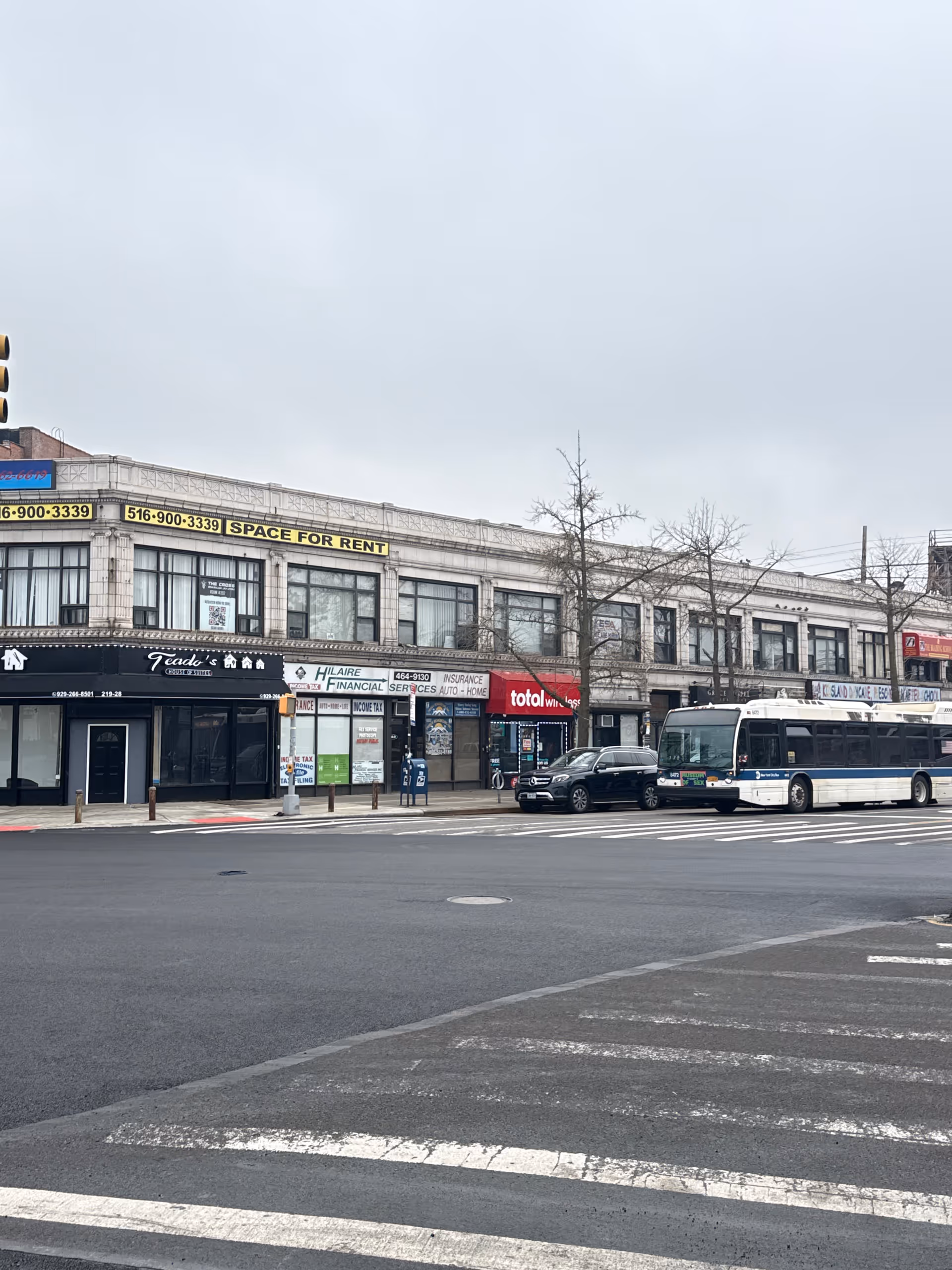 City street with storefronts, a parked bus, crosswalk, and overcast sky.