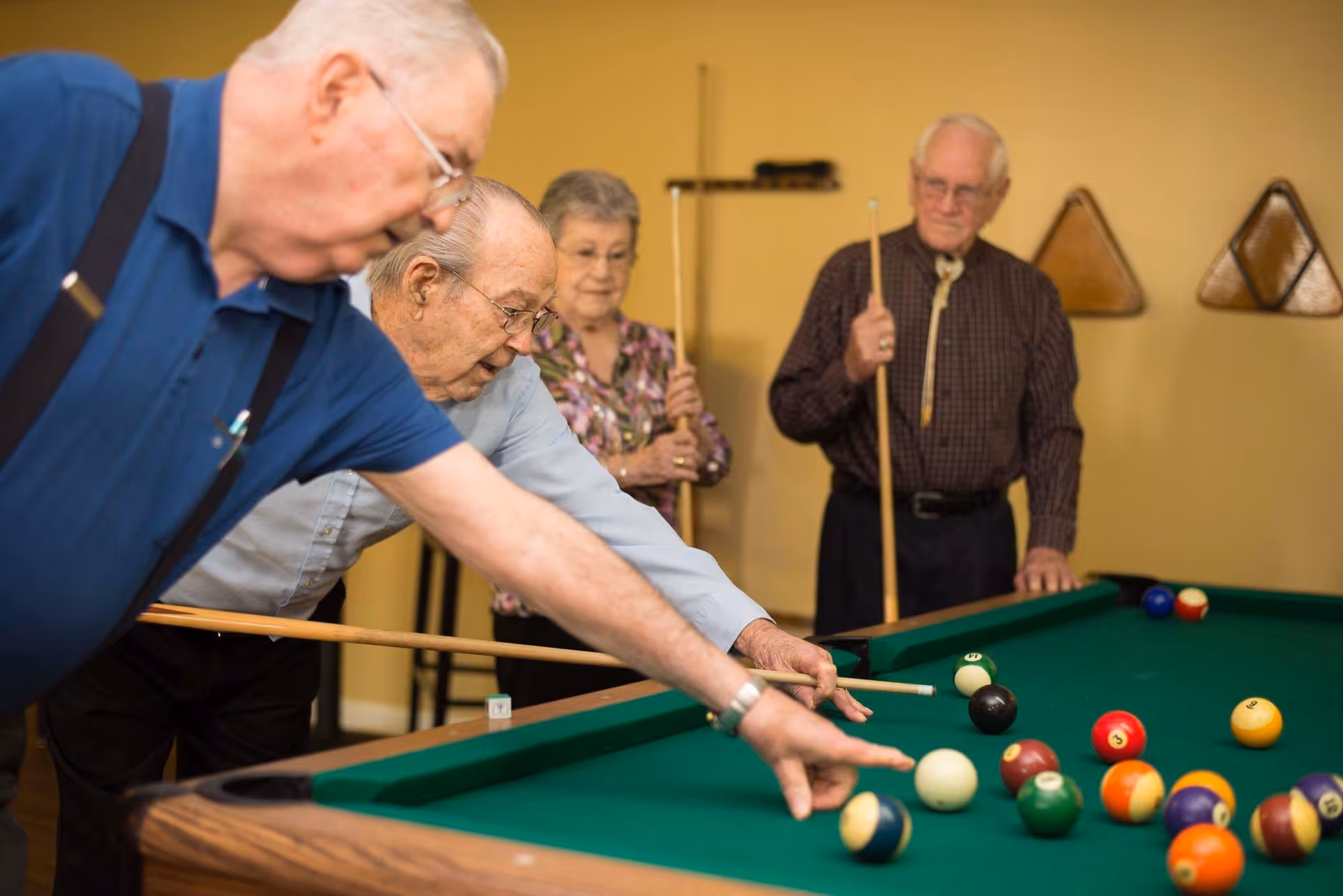 Four elderly individuals playing pool indoors. Two men are actively taking shots while a woman and another man stand nearby holding pool cues, watching the game. The room has a warm yellow wall with triangular wooden racks holding pool cues in the background.