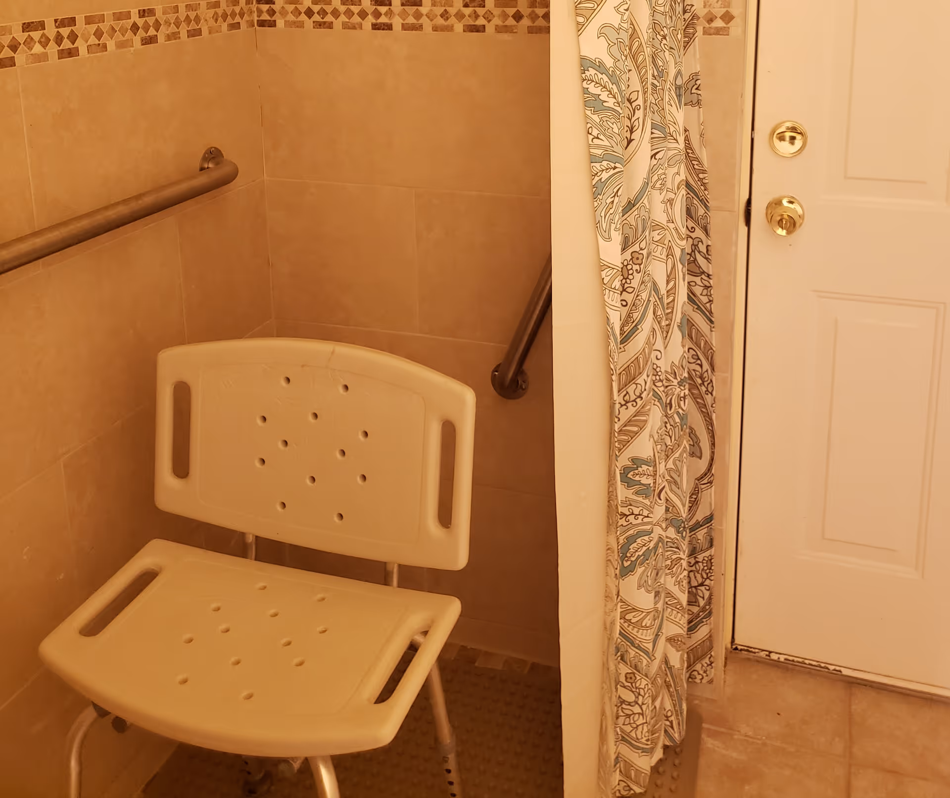 A beige tiled shower area with a white plastic shower chair and metal grab bars on the walls. A patterned shower curtain is partially visible next to a white door with gold-colored door handles.