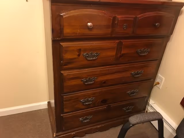 A tall wooden chest of drawers against a beige wall with a chair and an electrical outlet nearby.