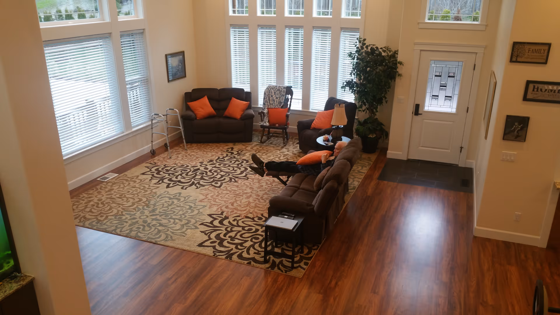 A cozy living room with large windows letting in natural light. The room features a patterned area rug, a brown sofa with orange pillows, a loveseat, a rocking chair, a side table with a lamp, and a walker near the window. There is a person reclining on the sofa. The entrance door has decorative glass panels, and there are framed pictures and signs on the walls.