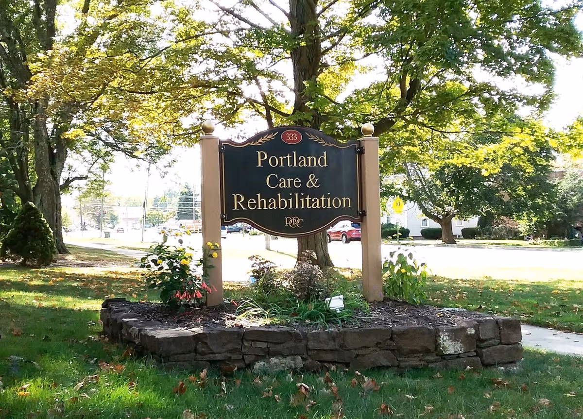 Outdoor view of a sign for Portland Care & Rehabilitation Centre Inc, mounted on two wooden posts surrounded by a stone border with plants and trees in the background.