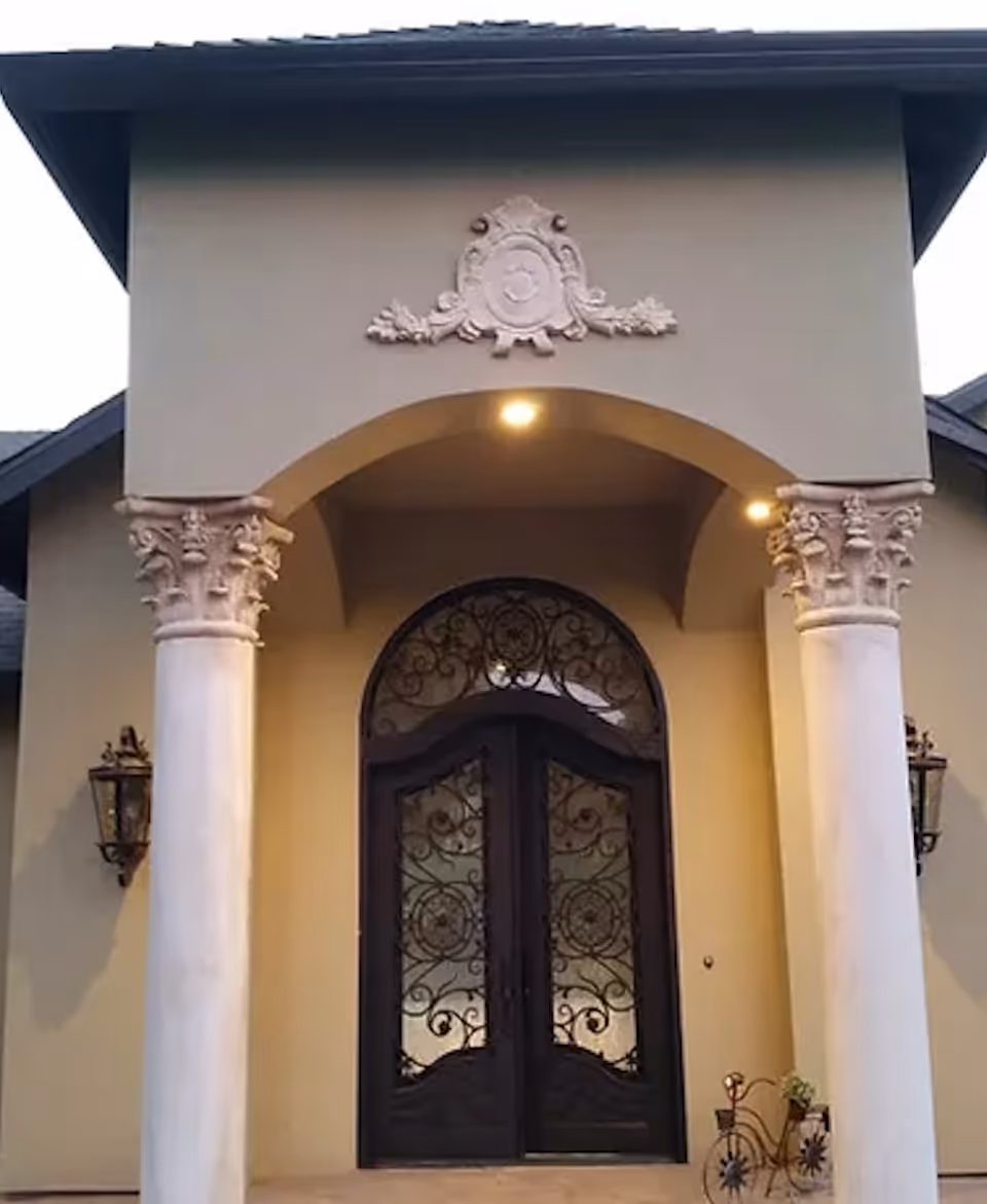 Arched front entrance with decorative columns and ornate double iron doors under a lit portico.