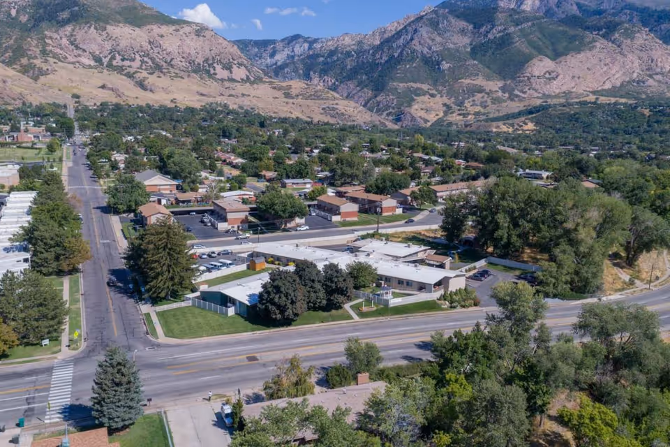 Aerial view of a single-story care facility and surrounding neighborhood with roads, trees, and mountains in the background.