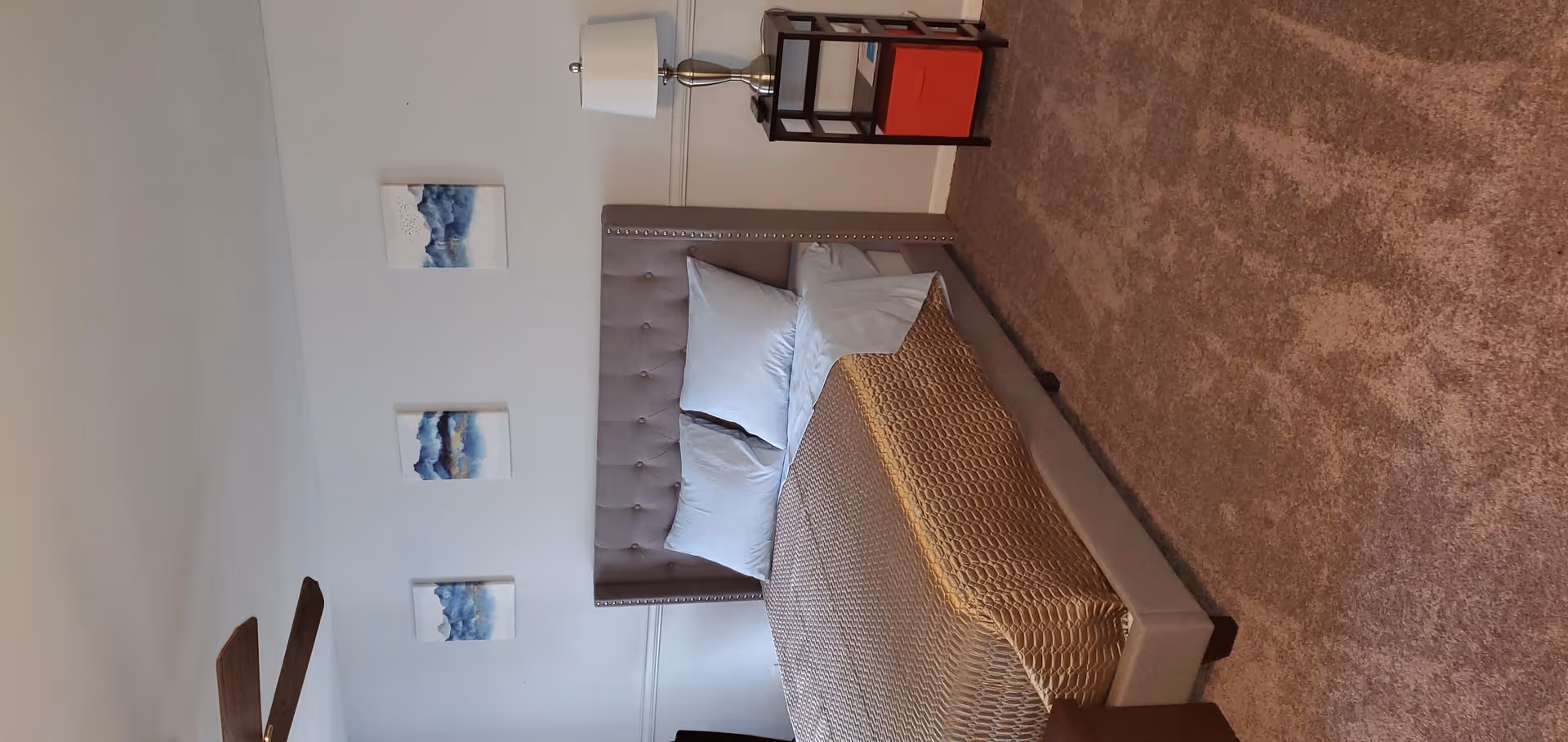 A carpeted bedroom with a tufted headboard bed made with pillows and a beige textured blanket, a nightstand with a lamp and shelving, and three small blue abstract wall artworks.