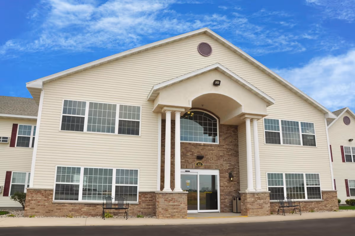 Front exterior view of Twin Town Villa Assisted Living Community building with beige siding, brick accents, large windows, and a covered entrance supported by four white columns under a blue sky with some clouds.