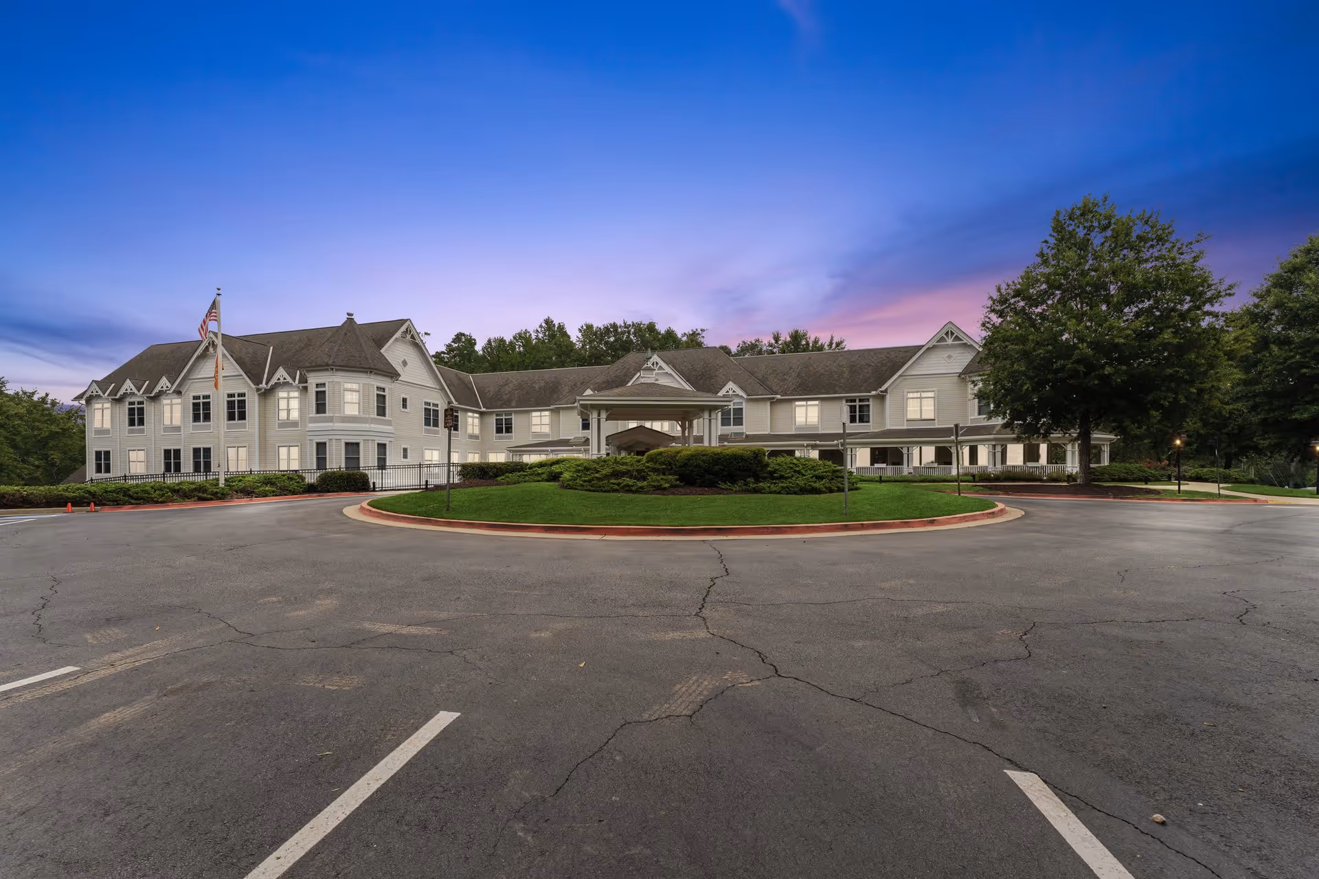 Exterior view of a large, two-story assisted living facility building named Assisted Living-Five Forks at dusk, with a circular driveway and well-maintained landscaping including bushes and a large tree. The sky is a gradient of blue and purple as the sun sets.
