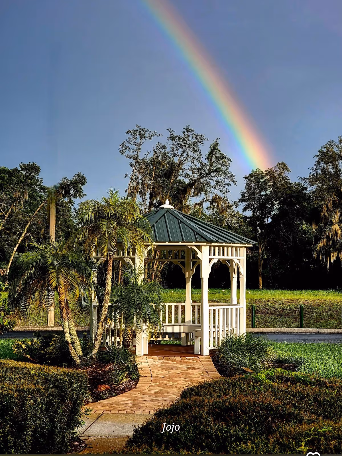 A white gazebo sits in a landscaped garden with palm trees and a rainbow overhead.