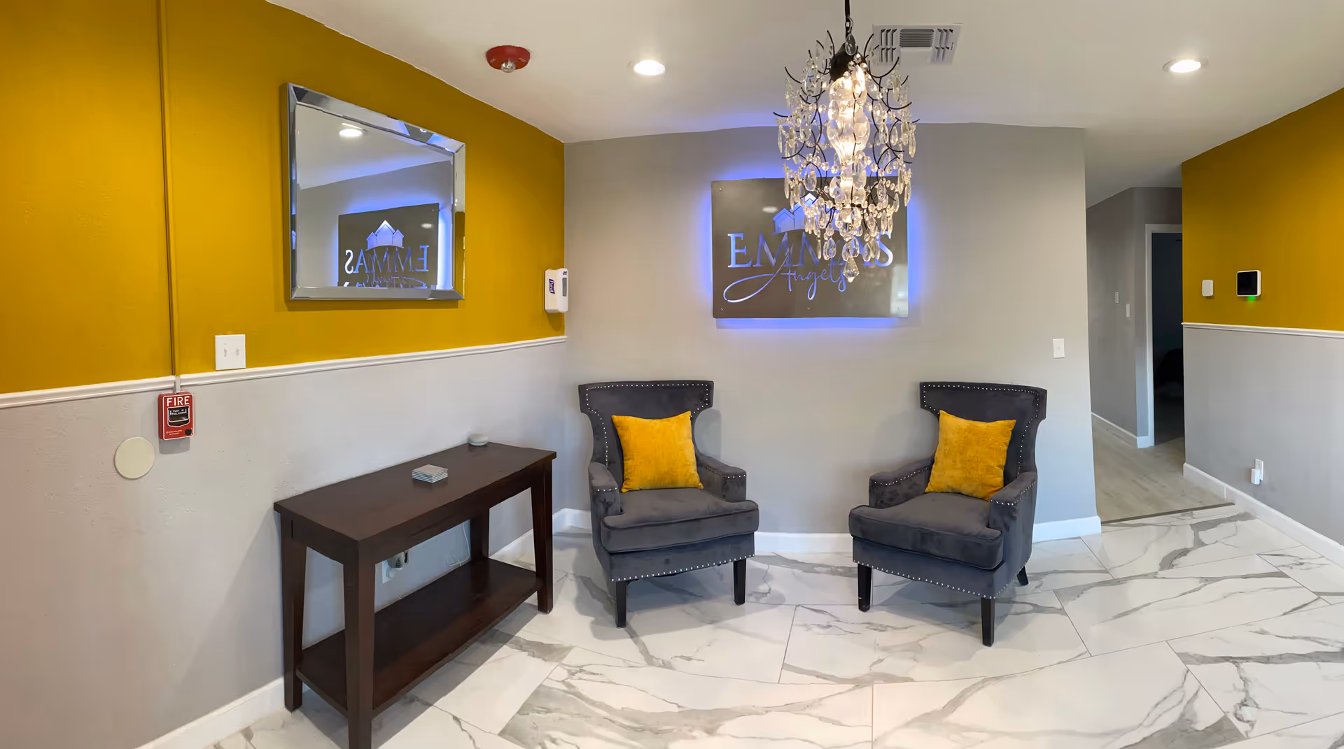 A modern facility lobby featuring two gray chairs, chandelier, yellow walls, and marble floor.