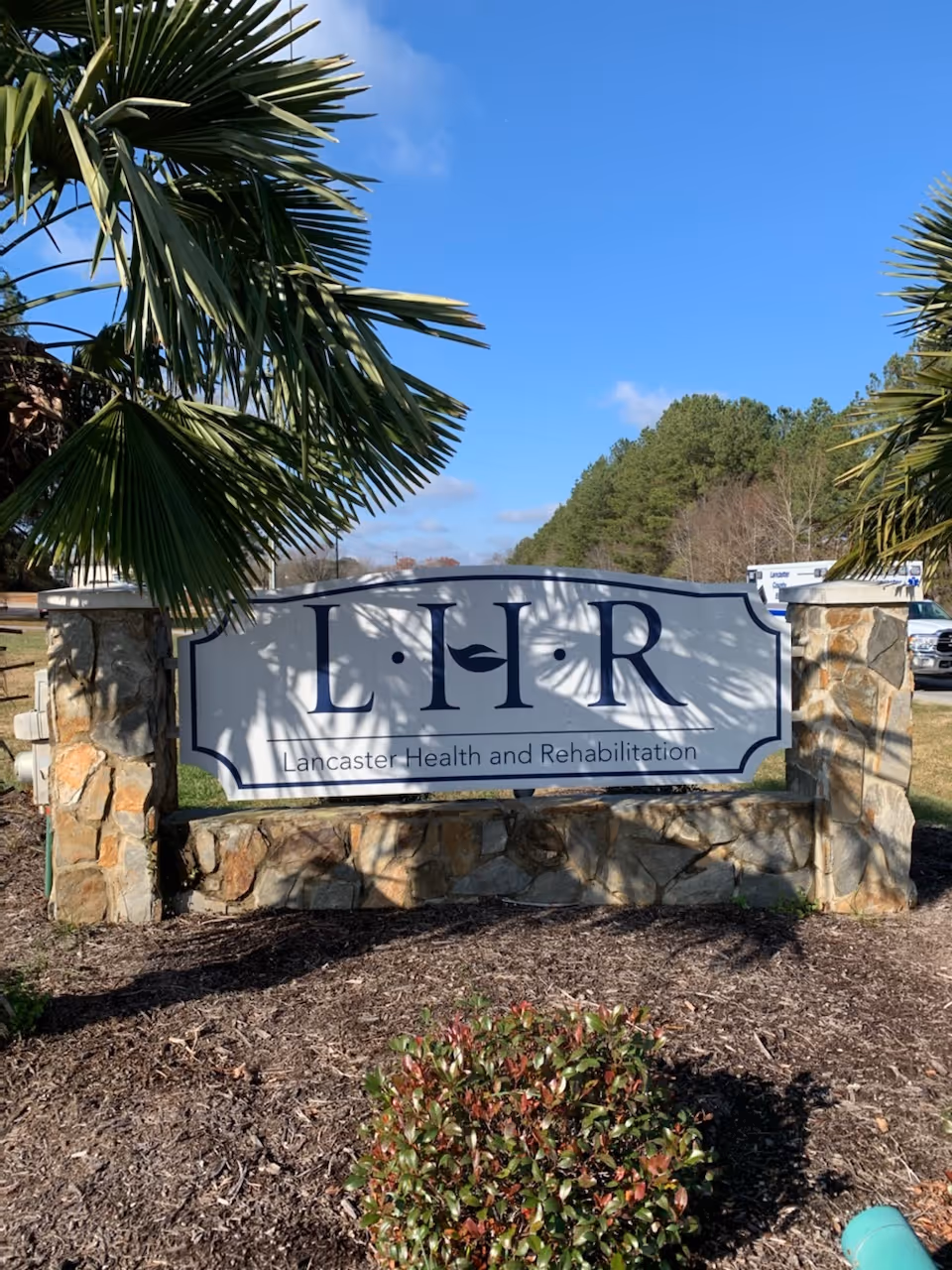 Stone sign for Lancaster Health and Rehabilitation outdoors with palm trees.