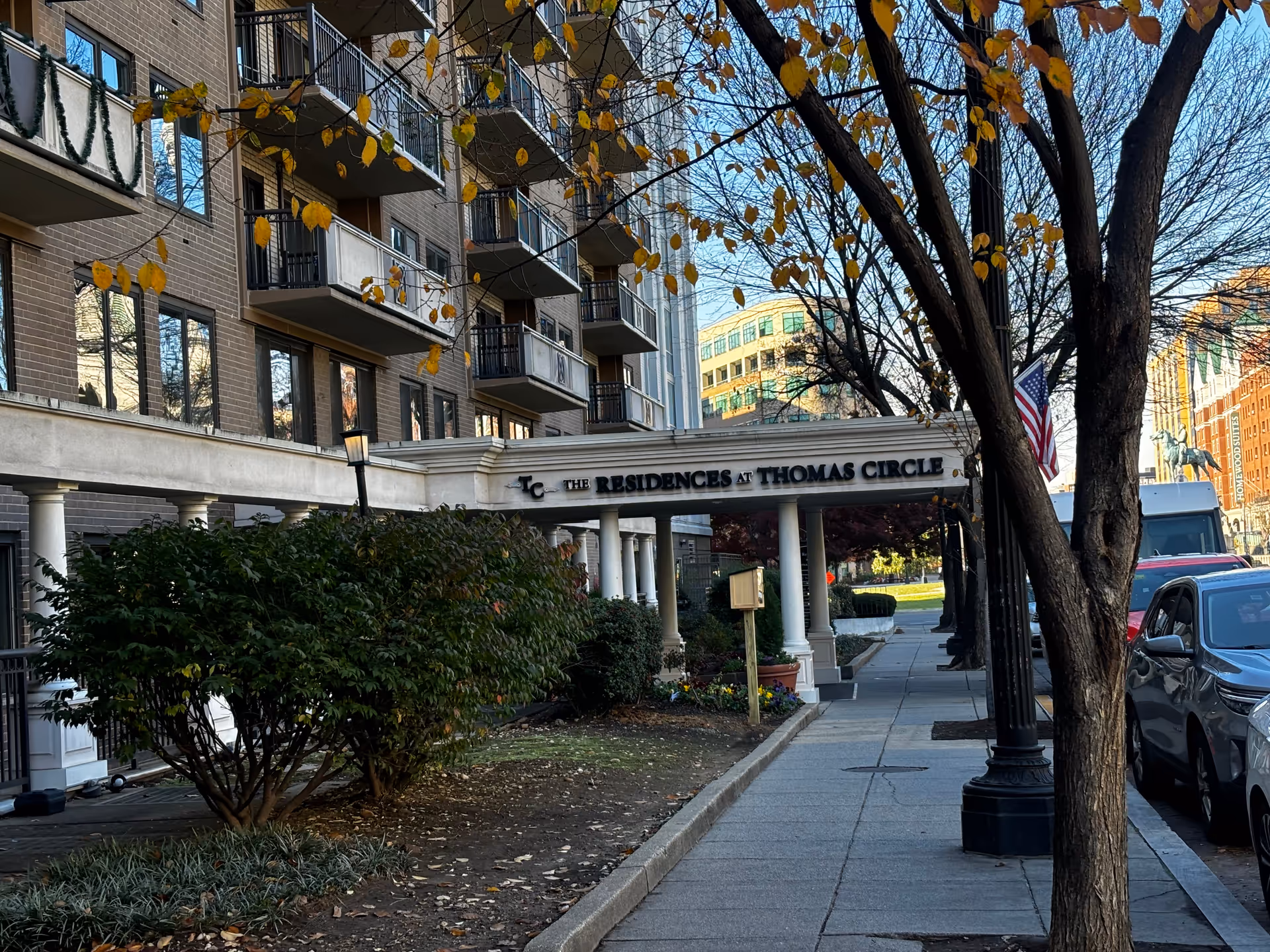 Front entrance of a multi-story brick residential building with a covered portico reading 'The Residences at Thomas Circle,' balconies, trees, and a sidewalk.