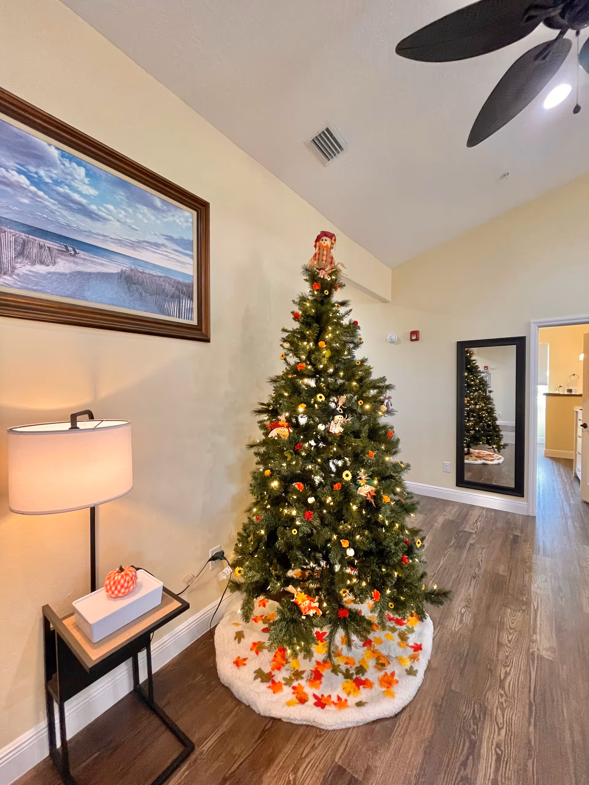 A decorated Christmas tree stands in a lit living room corner with a mirror and lamp.