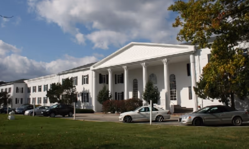 White, neoclassical hospice building with tall columns, a wide front entrance, and parked cars.