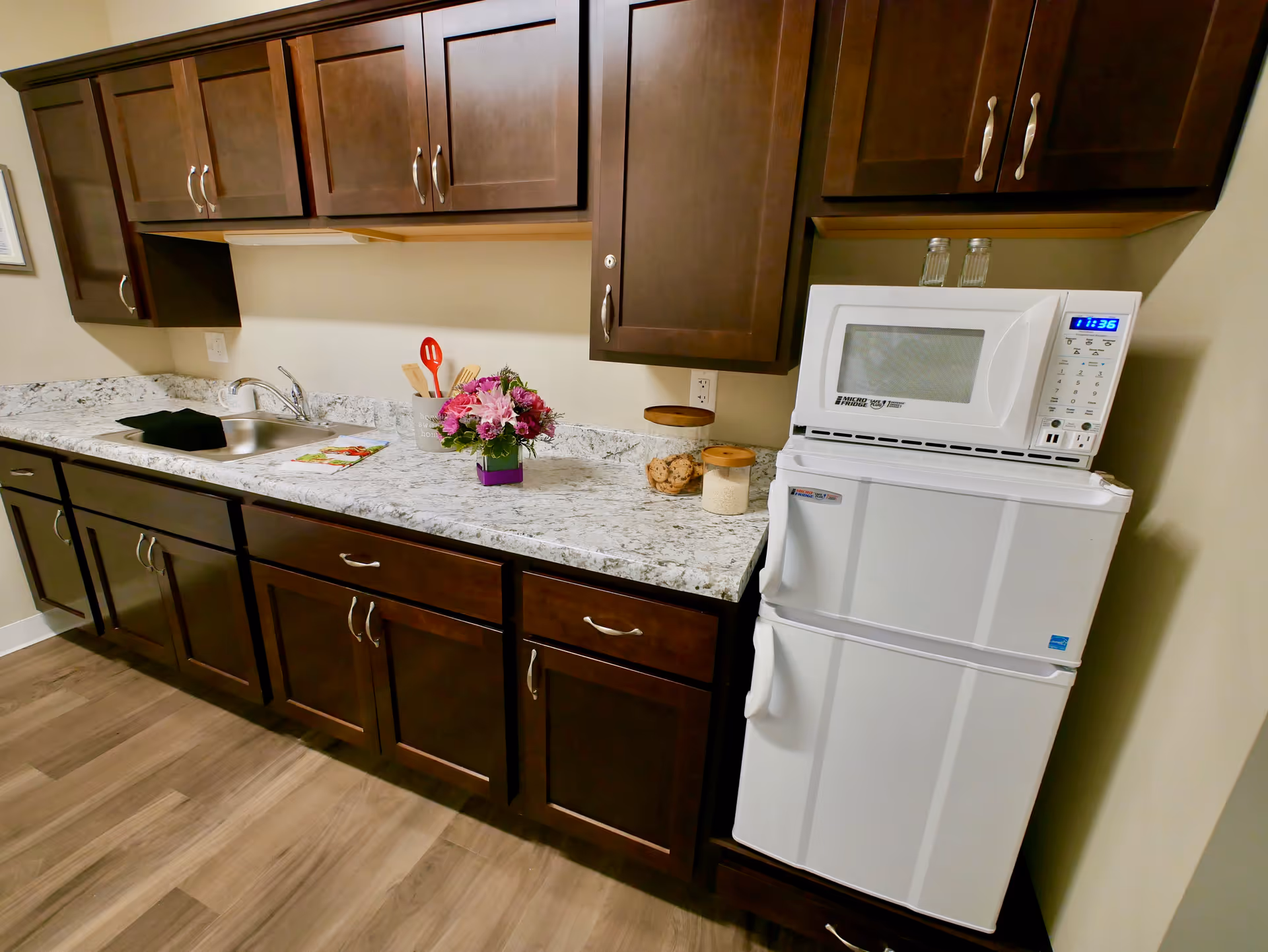 A kitchen area with dark wooden cabinets, a white marble countertop, a stainless steel sink, a white microwave on top of a white mini refrigerator, and various kitchen items including a flower vase, cooking utensils, and jars with cookies and grains.