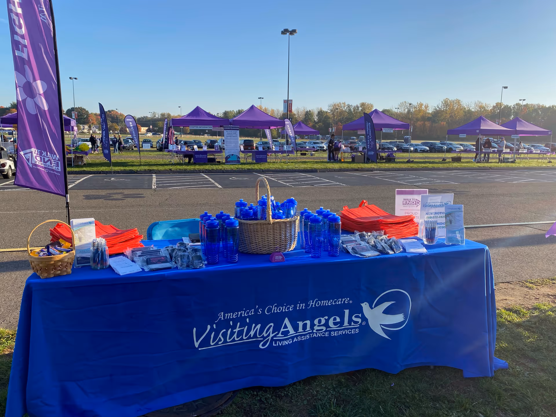 Promotional table with blue cover for Visiting Angels, set up outdoors with tents and signs.