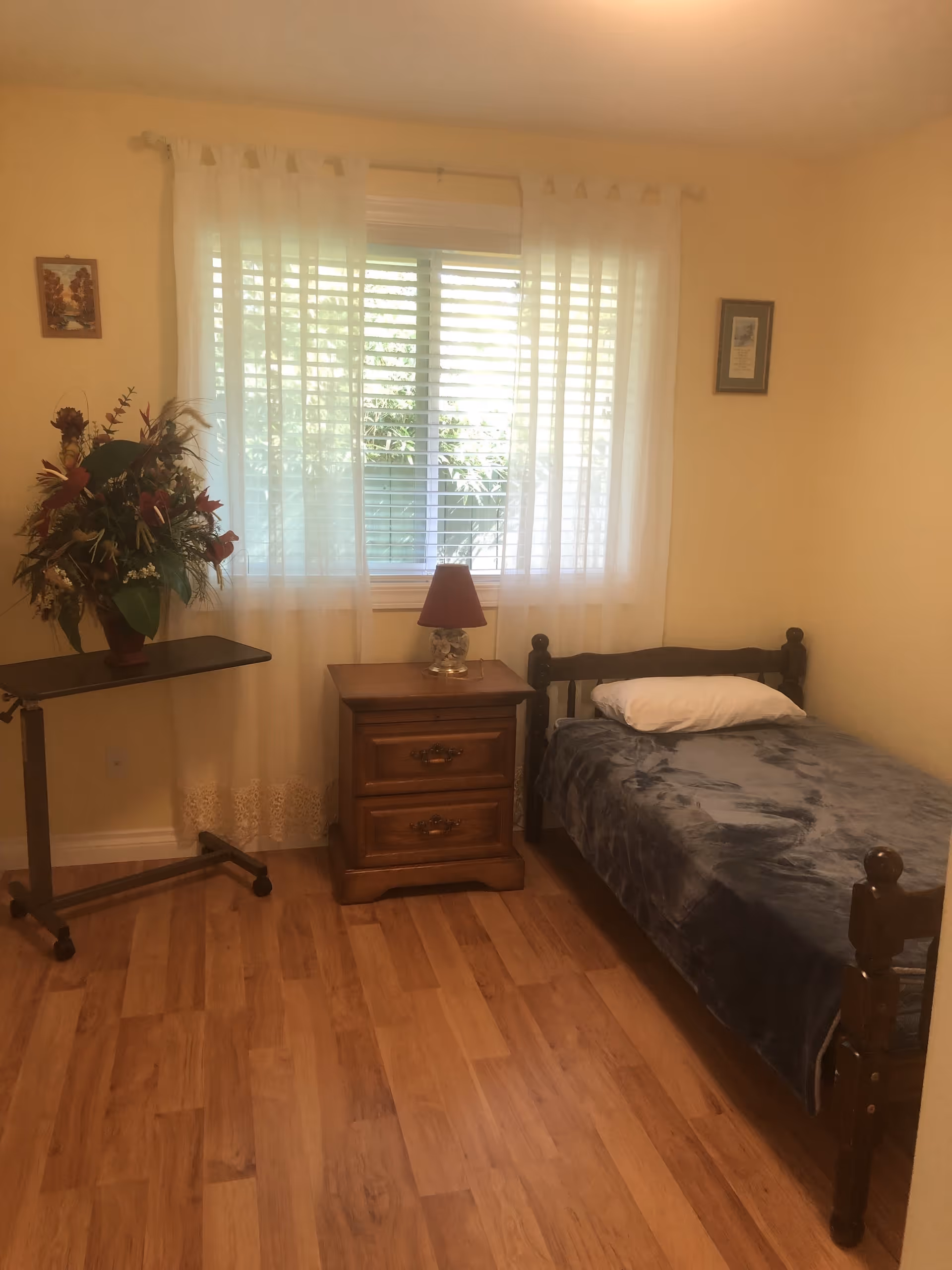 A small bedroom with a single wooden bed covered with a dark gray blanket and a white pillow. Next to the bed is a wooden nightstand with two drawers and a small lamp with a red shade. A window with white sheer curtains and blinds is centered on the wall behind the nightstand. To the left of the nightstand is a small rolling table with a large floral arrangement. The room has light yellow walls and wooden flooring.