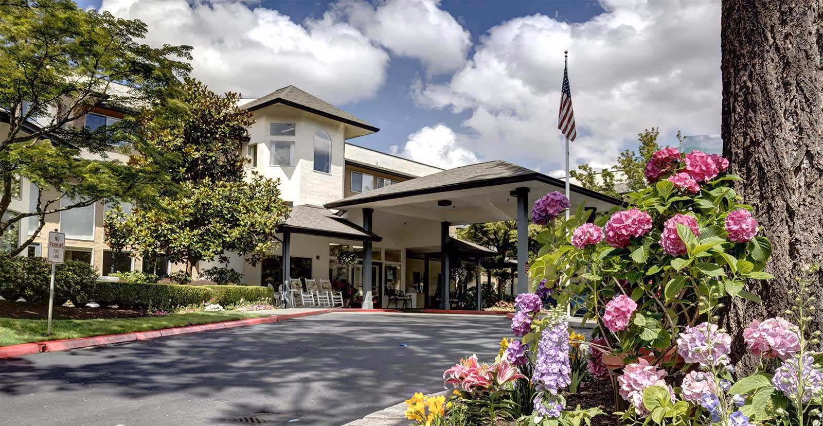 Exterior view of a senior living facility with a covered entrance, surrounded by trees and colorful flowers in the foreground under a partly cloudy sky.