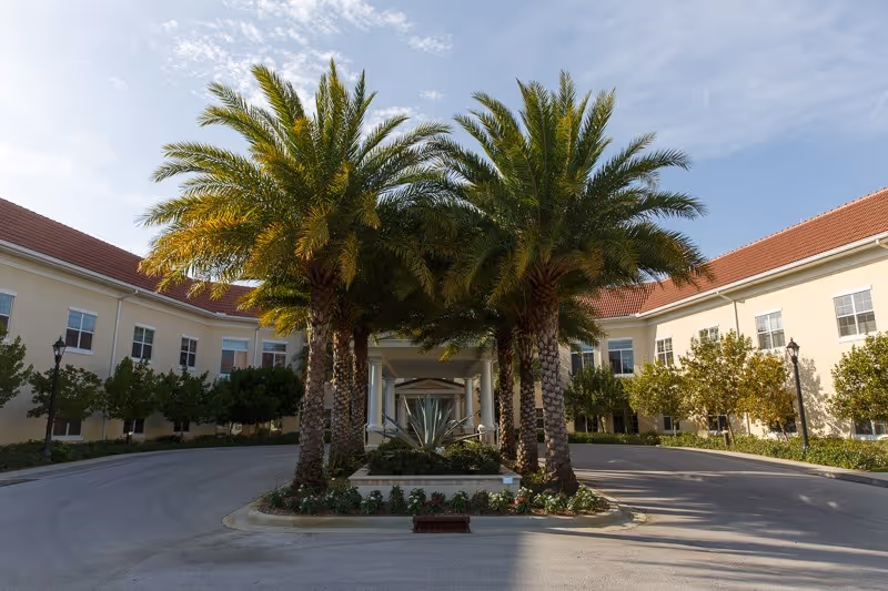 Entrance driveway of a senior living facility with a circular driveway lined by palm trees and landscaped plants, flanked by two beige buildings with red-tiled roofs under a partly cloudy sky.