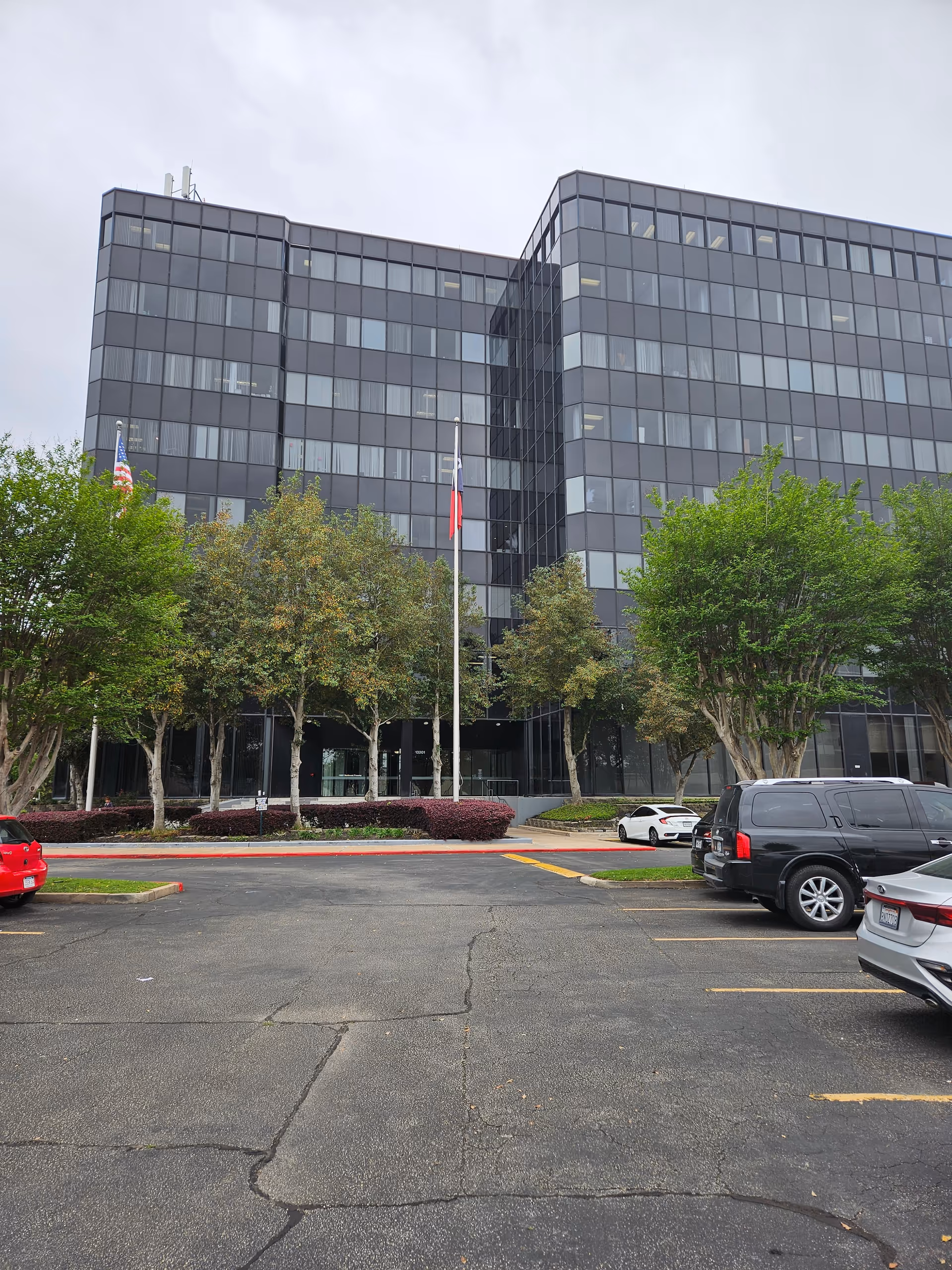 Front view of a modern glass office building with trees, flags, and a parking lot.