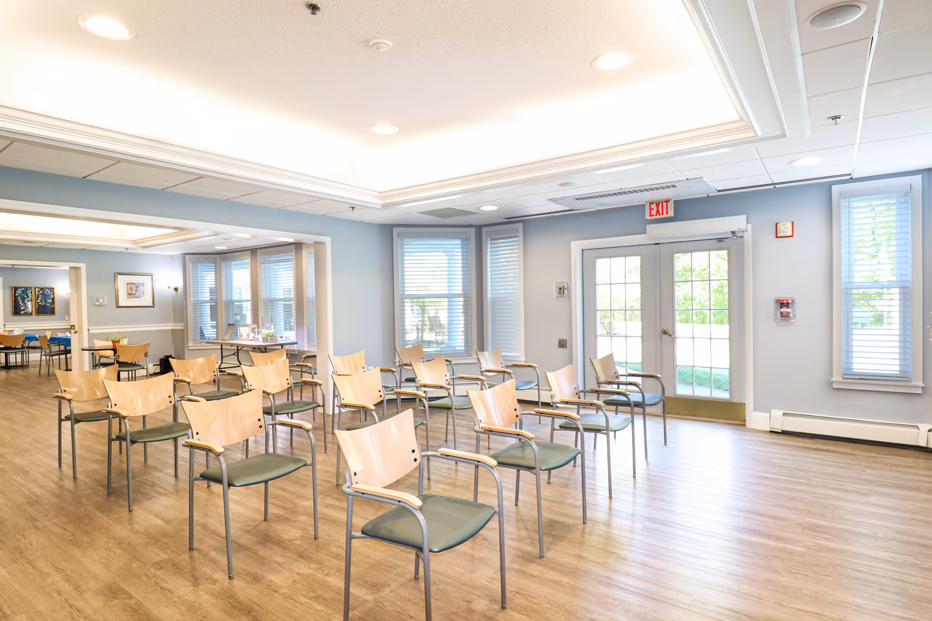 Empty community room with rows of wooden-backed chairs and large windows.