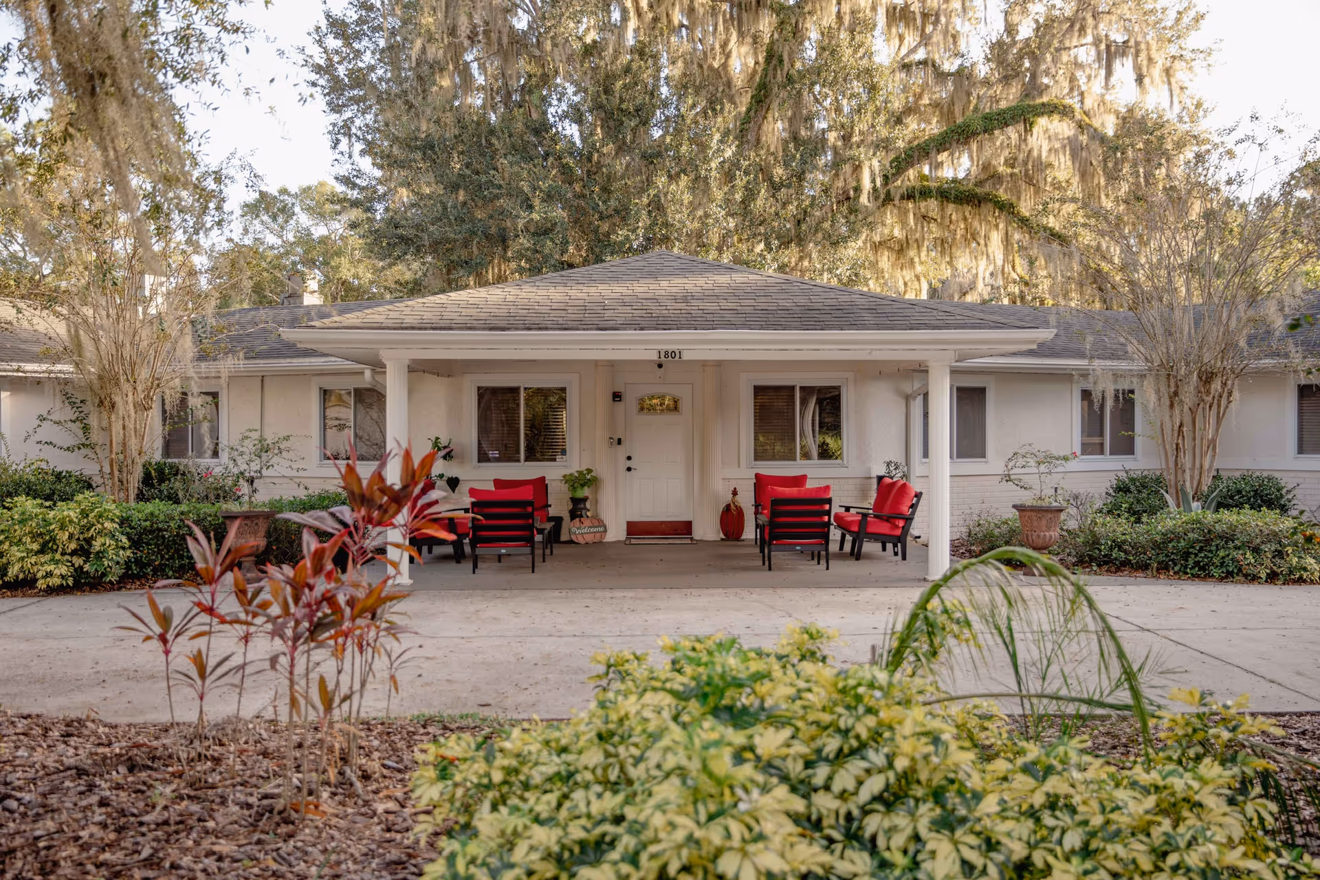 A single-story white senior living building with a covered front porch and red chairs.