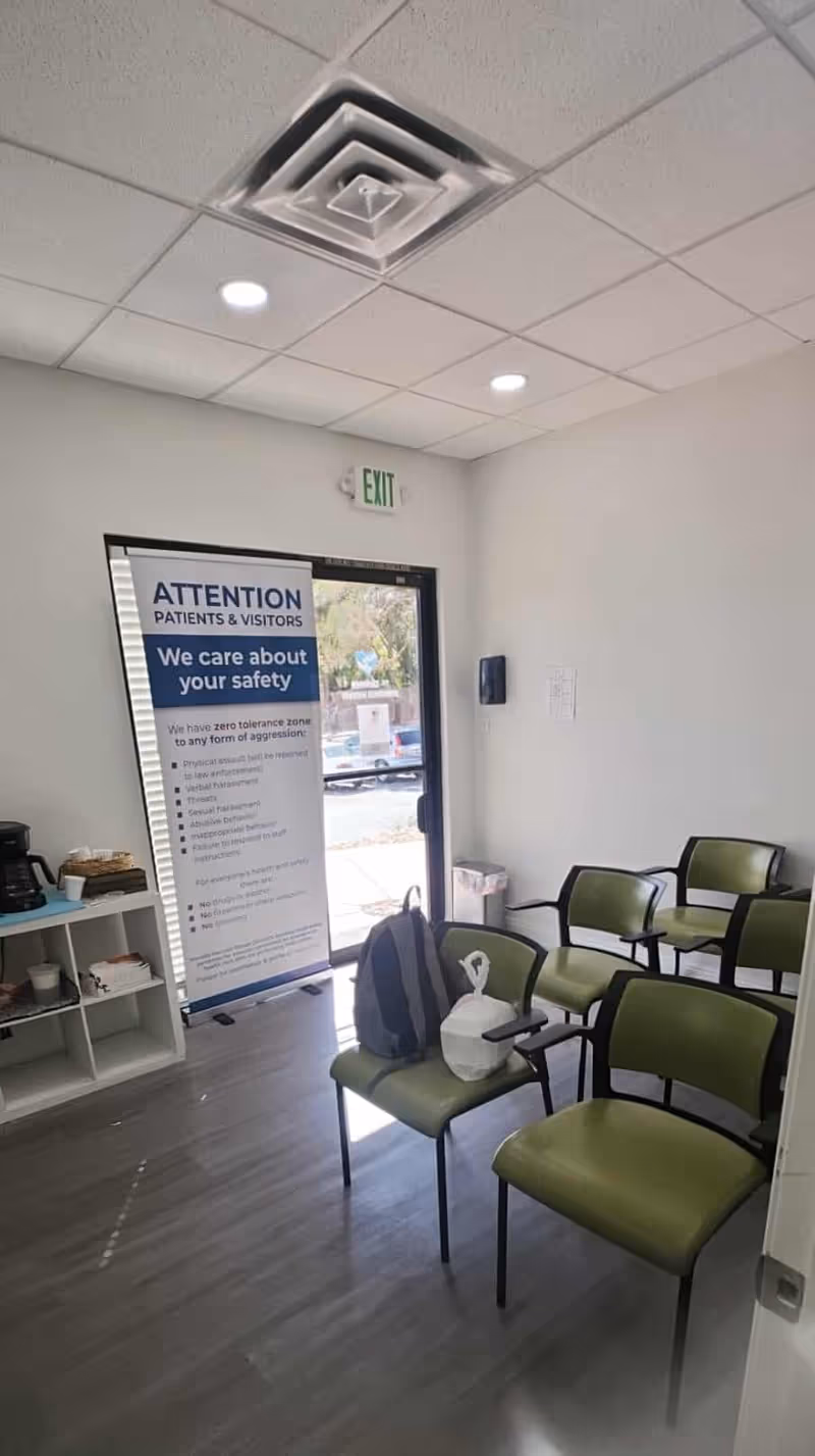 Interior waiting area with green chairs, an exit sign, banner, door, and shelves.