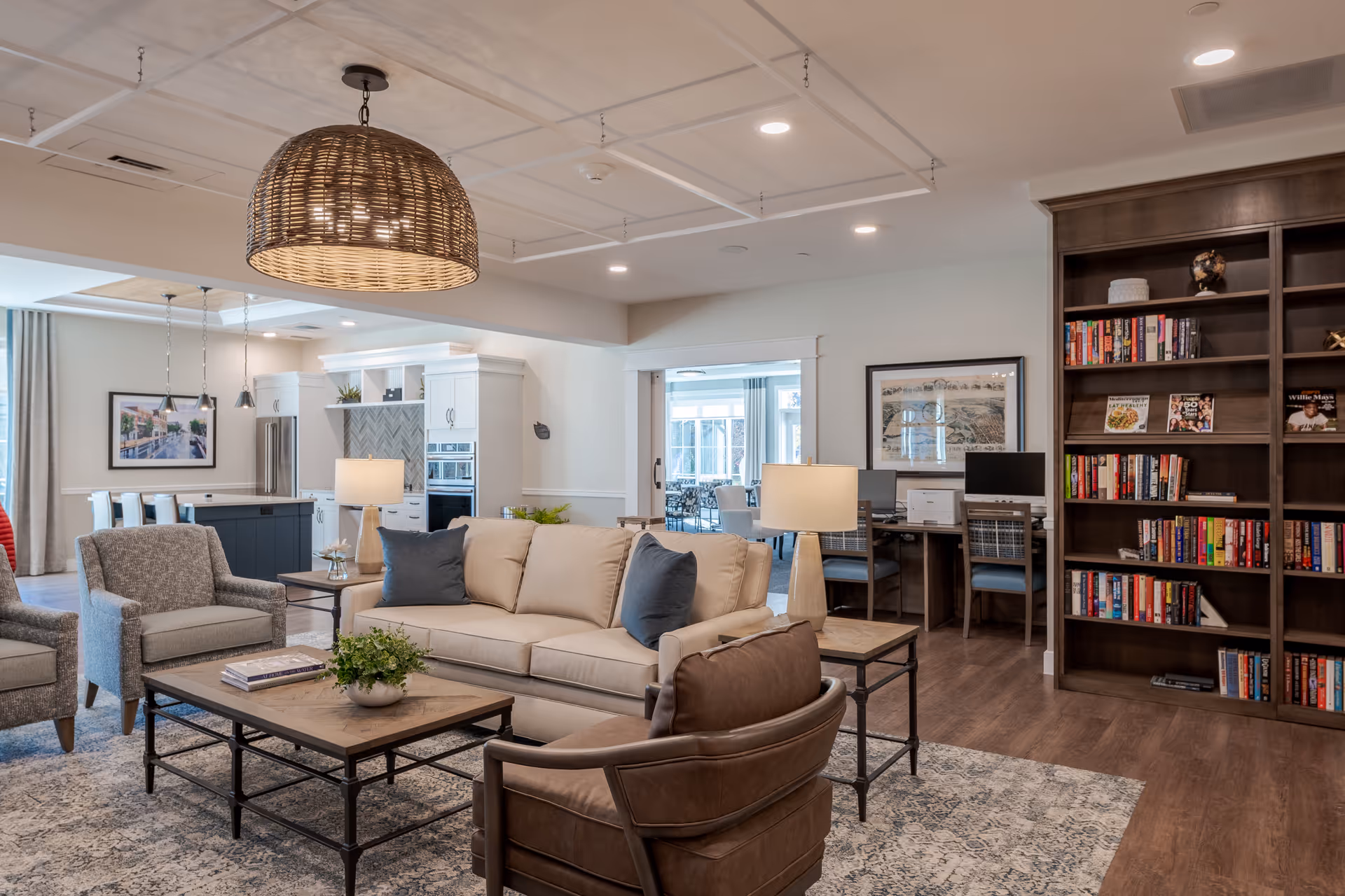 Cozy communal living room with sofas, armchairs, coffee tables, a large wicker pendant light, a bookshelf and an open kitchen area in the background.