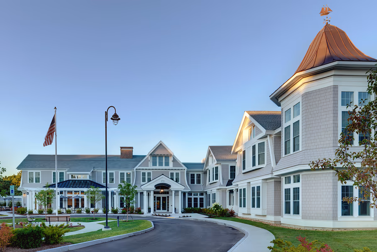 Exterior front entrance of a senior living residence with a circular driveway, flagpole, and landscaped grounds.