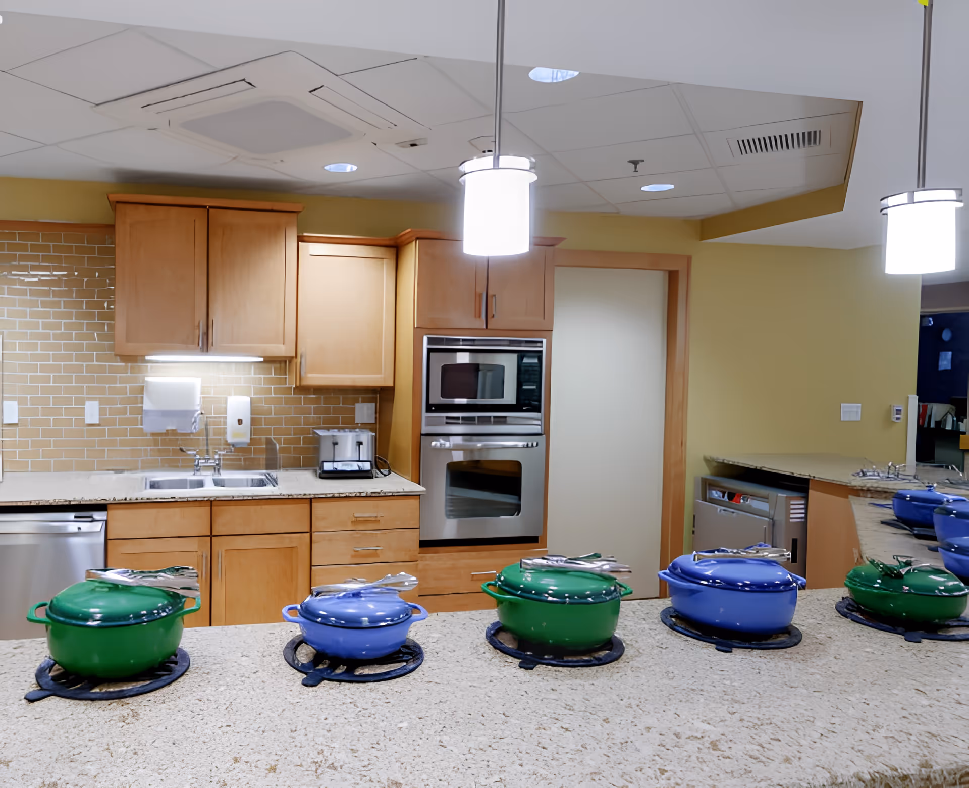 A modern kitchen area with light wood cabinets, a built-in oven and microwave, a stainless steel dishwasher, and a sink. On the countertop in the foreground, there are alternating green and blue covered pots with utensils placed on top, arranged in a row. The kitchen has a beige tiled backsplash and pendant lights hanging from the ceiling.