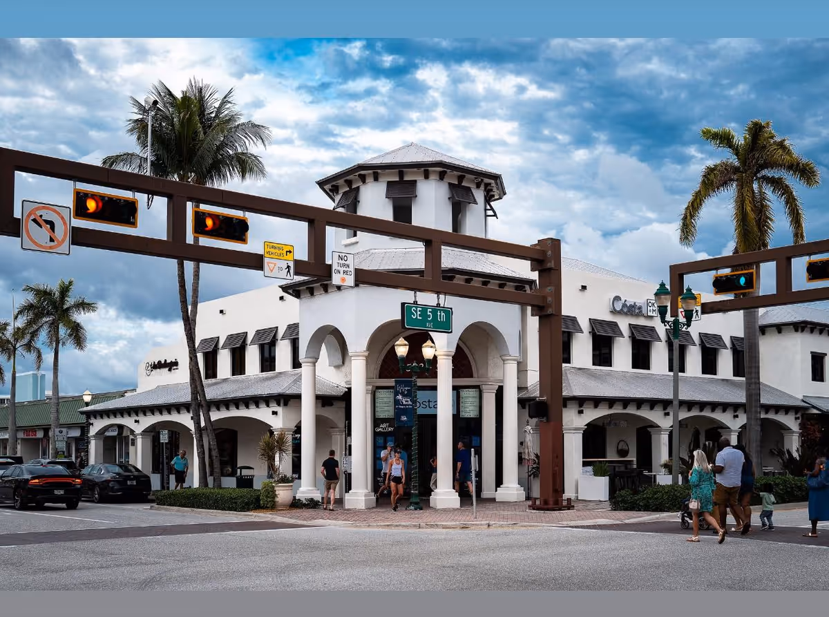 White colonial-style building with arches, palm trees, and pedestrians along a street.