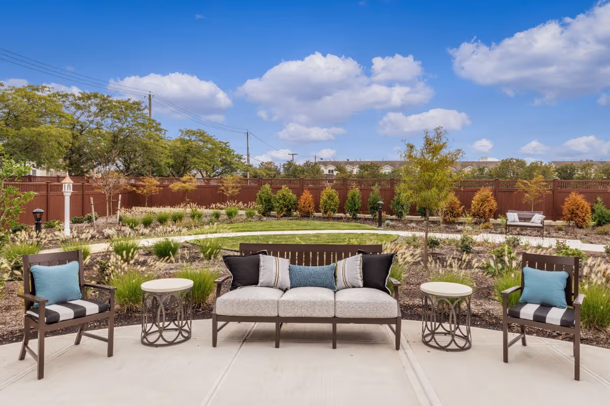 Outdoor courtyard with a sofa and chairs on a patio facing a landscaped garden and wooden fence under a blue sky.