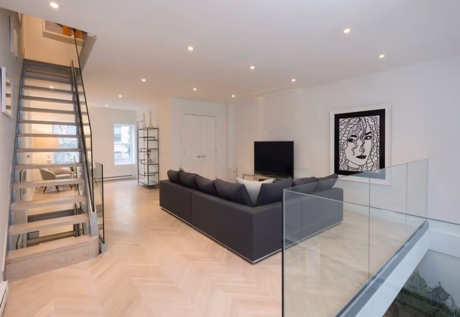 Modern open-concept living area with gray sectional, TV, staircase, and glass railing.