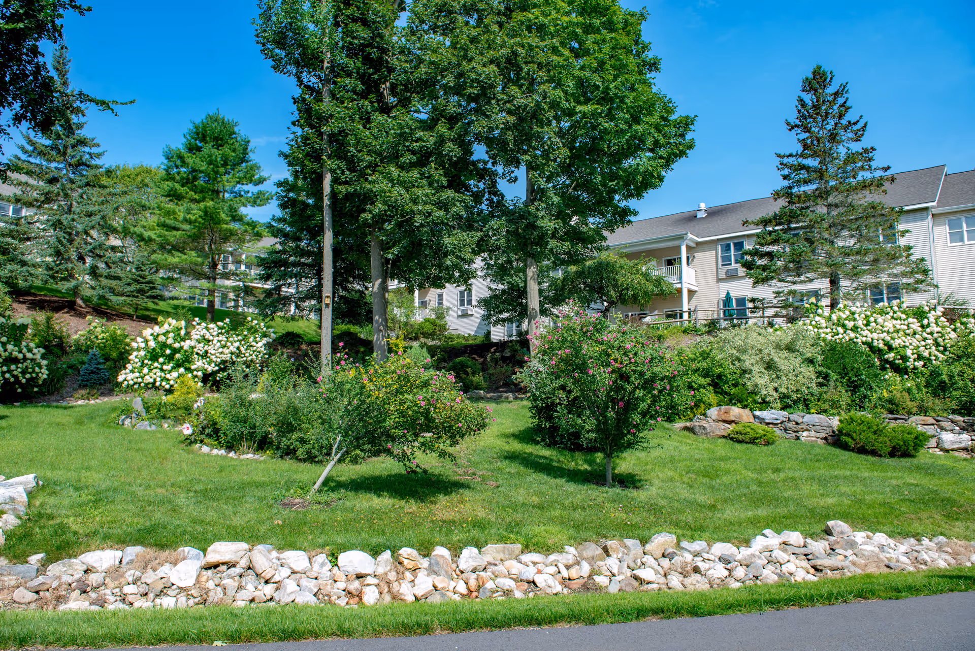 A landscaped outdoor area with green grass, various trees, and flowering bushes in front of a multi-story residential building under a clear blue sky.