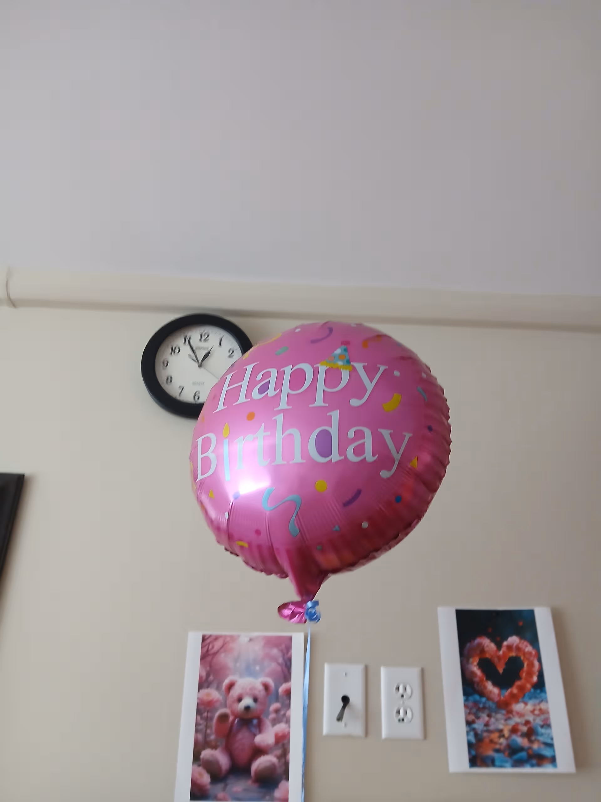 Pink Happy Birthday balloon near a wall clock with framed pictures indoors.
