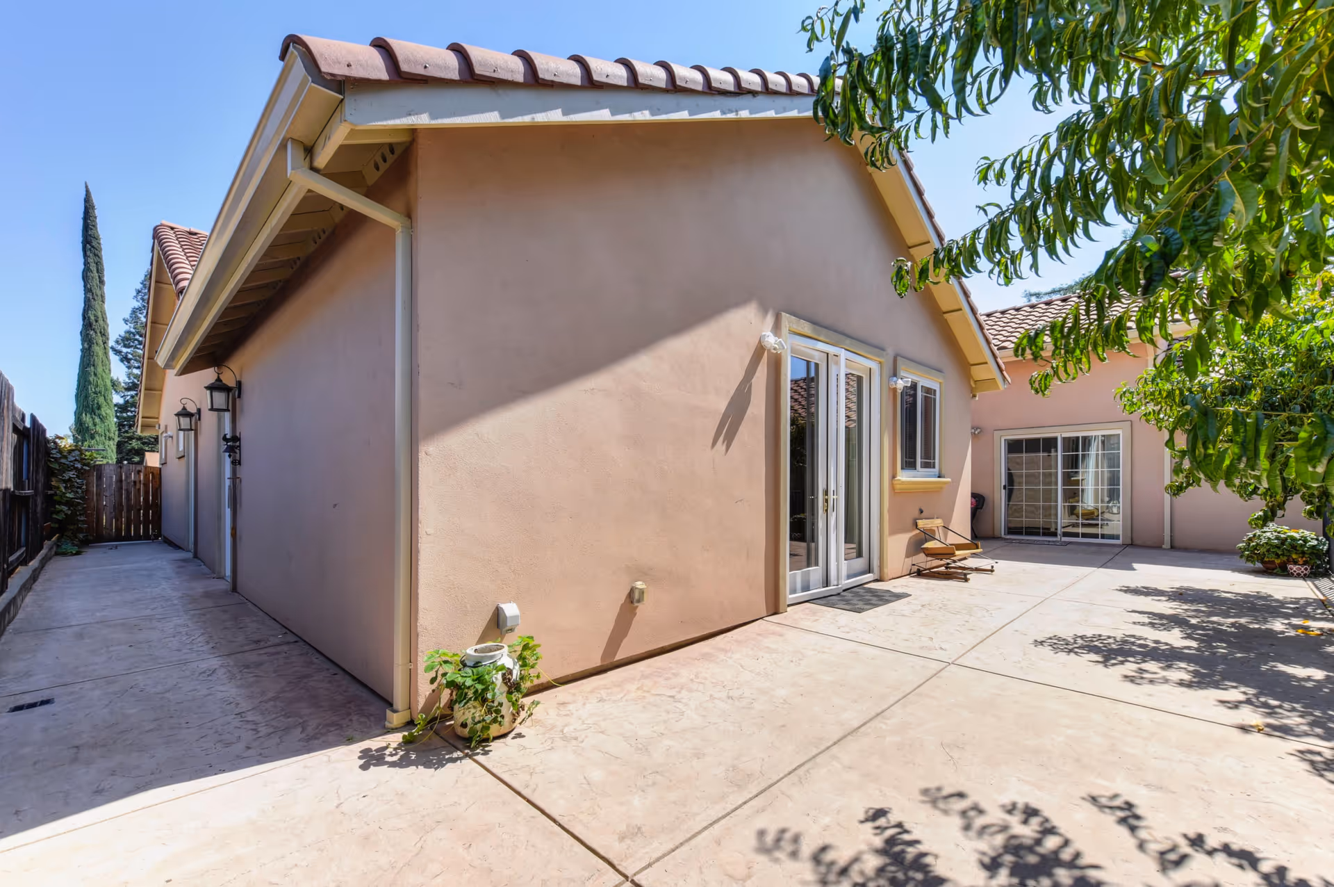 Sunlit paved courtyard beside a single-story stucco building with French doors, a sliding glass door, and nearby trees.