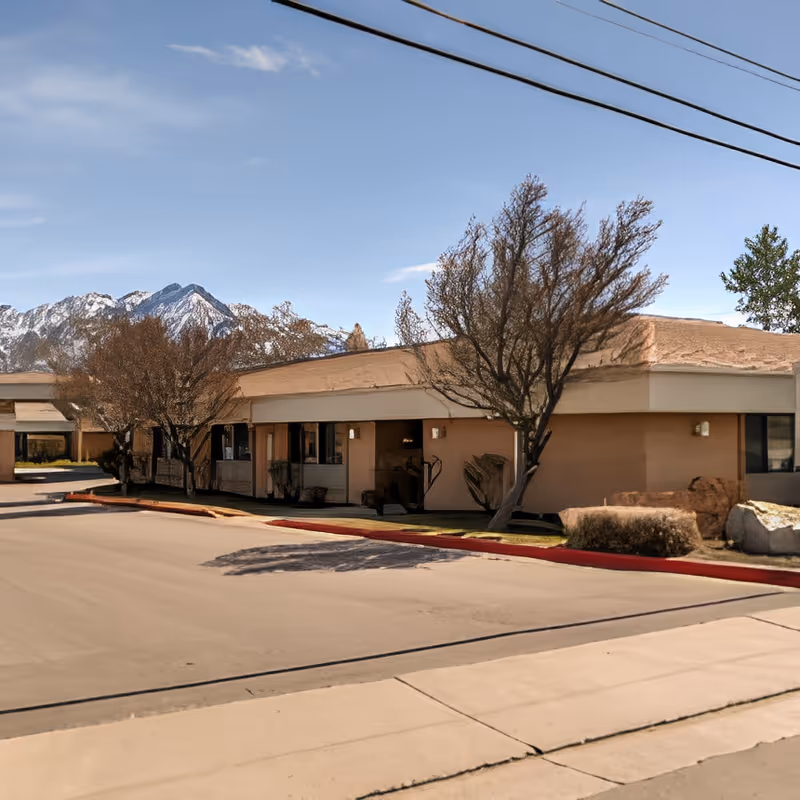 Single-story senior living facility front with trees, a driveway, and snow-capped mountains in the background.
