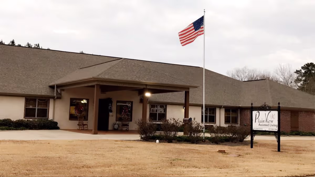 Exterior view of Plain View Assisted Living facility showing a single-story building with a covered entrance, an American flag on a flagpole, and a sign with the facility's name in front on a lawn.