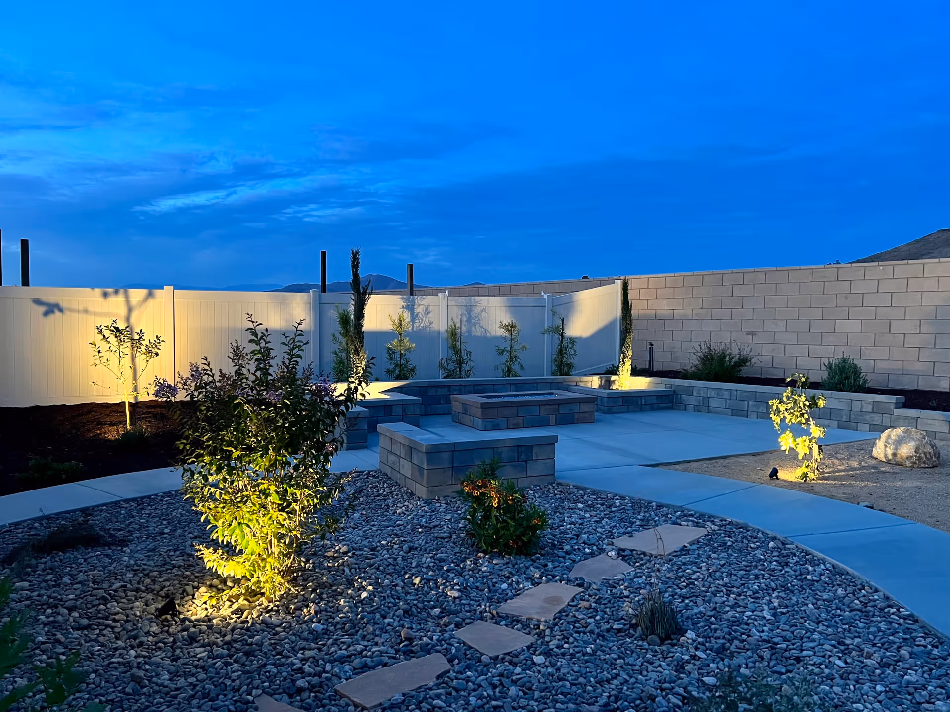 Outdoor patio area at dusk with stone benches and a fire pit surrounded by a landscaped garden with small plants and trees, enclosed by a white fence and a brick wall.