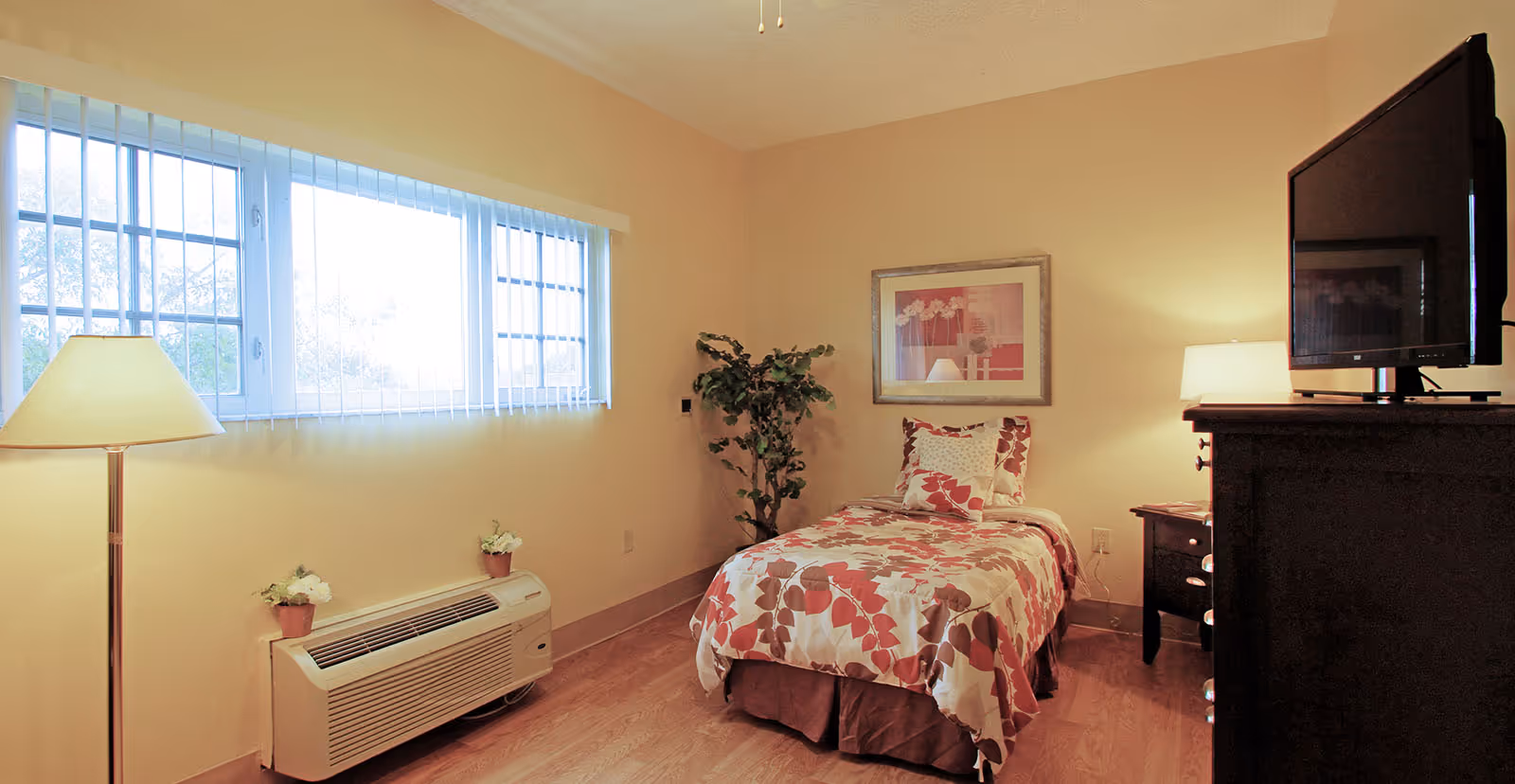 A small bedroom with a single bed covered in a floral-patterned bedspread and pillows. The room has light-colored walls, a large window with vertical blinds, a floor lamp, a potted plant in the corner, a wooden dresser with a flat-screen TV on top, and a nightstand with a table lamp.