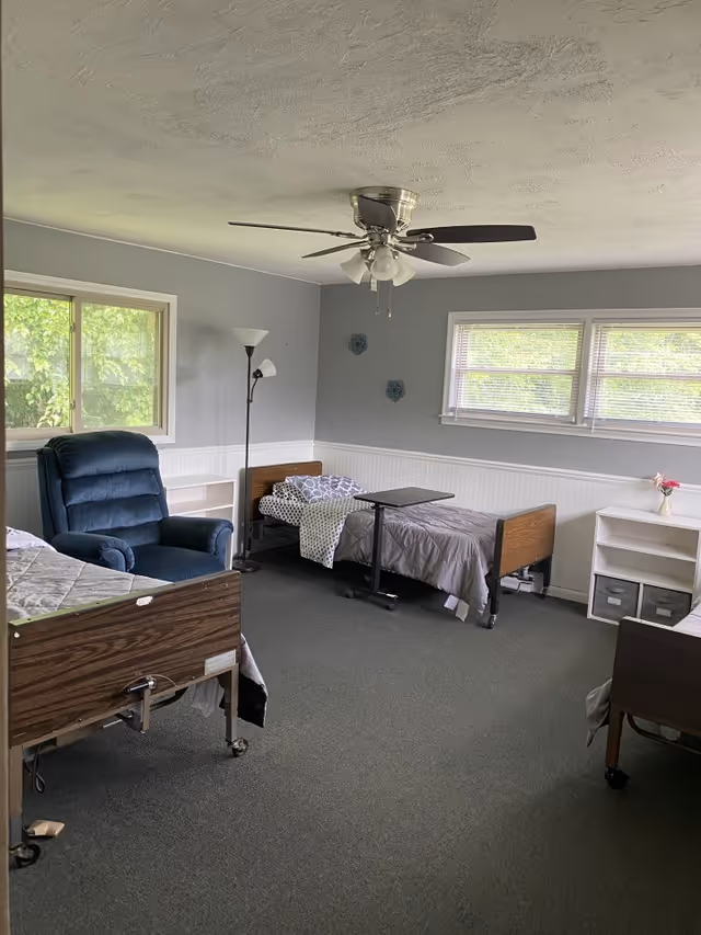 A senior living facility bedroom with two hospital-style beds, a blue recliner chair, a floor lamp, a ceiling fan with lights, and windows letting in natural light. The walls are painted gray with white wainscoting, and there are small decorative items on the wall and a white shelving unit with storage bins.