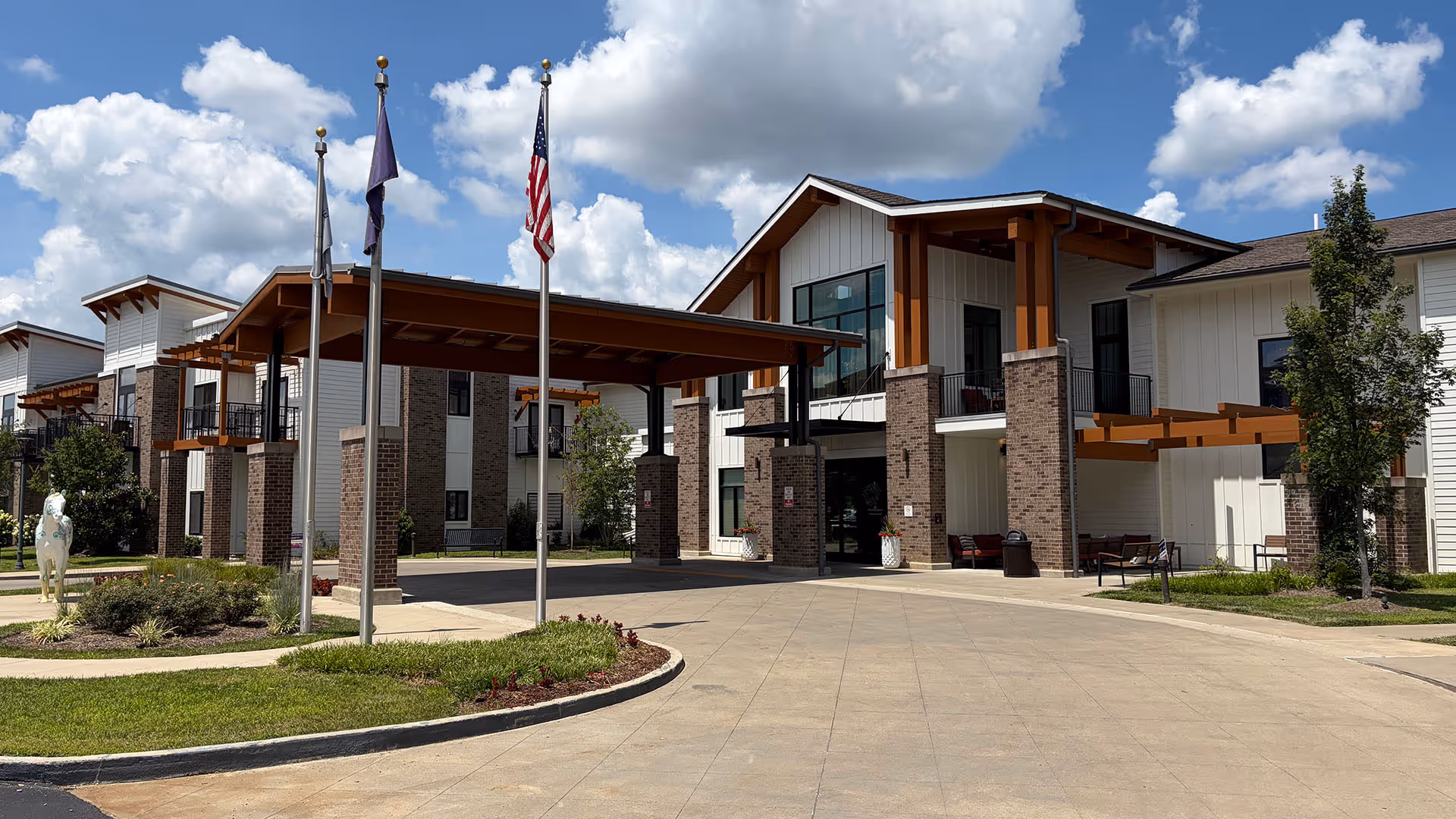 Modern senior living facility exterior with brick pillars, a porte-cochère, and flagpoles.