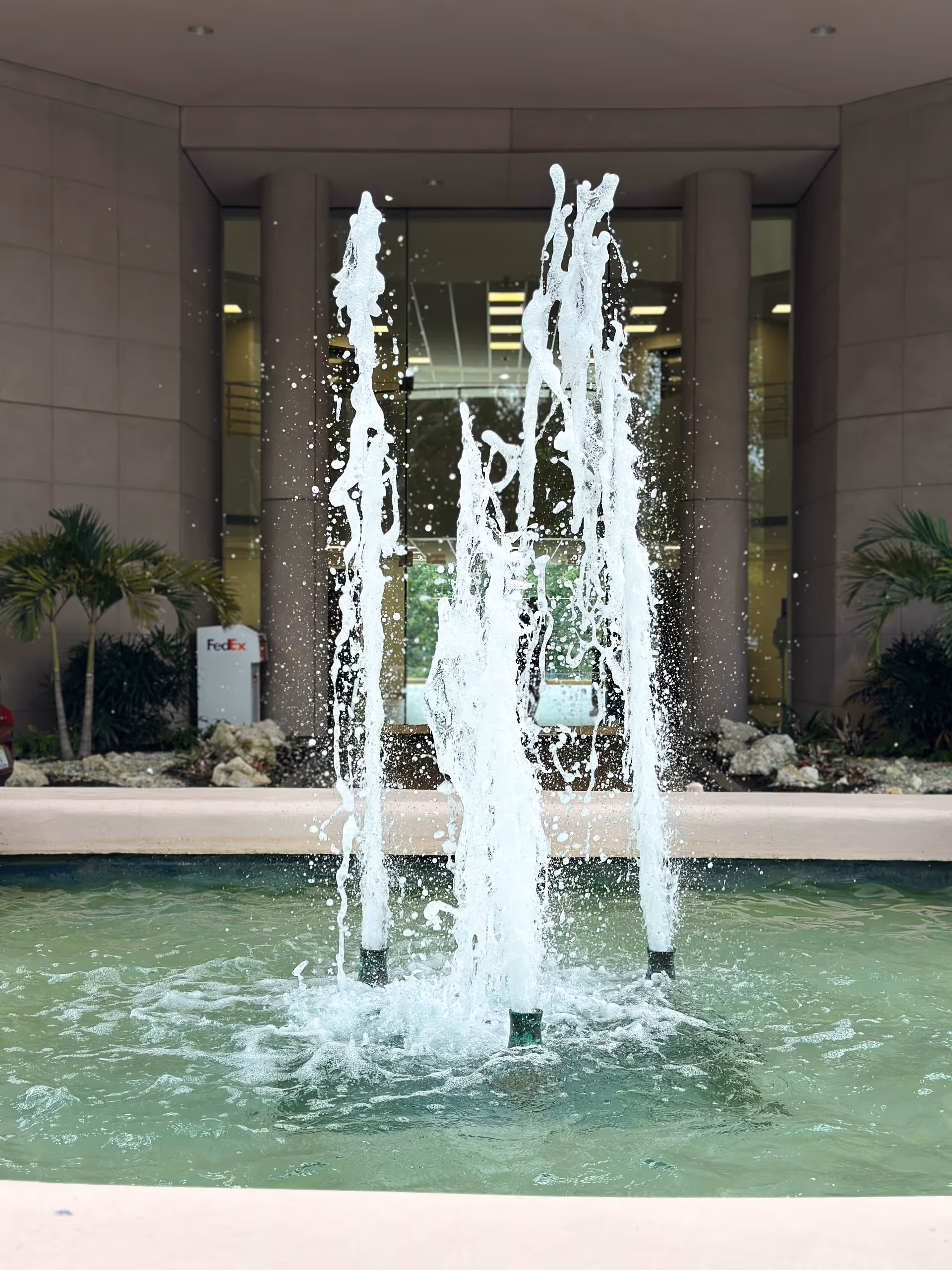 Water fountain jets shoot upward in a pool at the front entrance of a modern building.