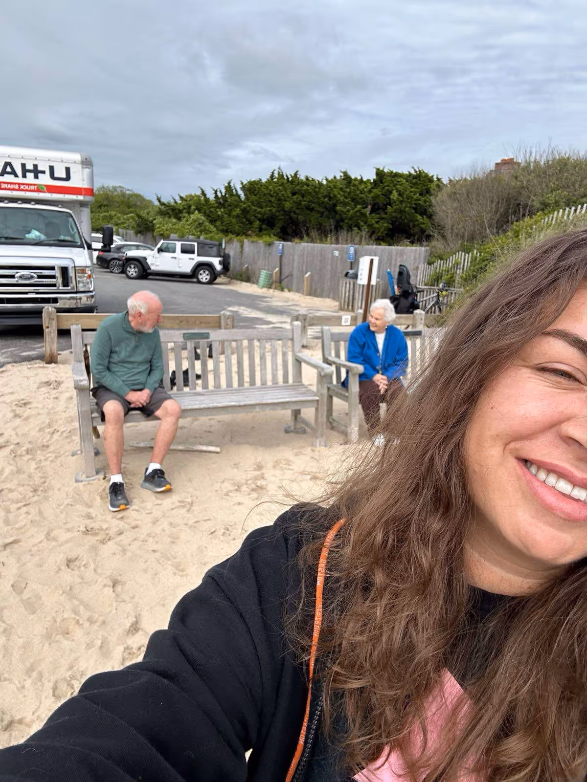 A smiling woman takes a selfie on a sandy outdoor seating area with two elderly residents.