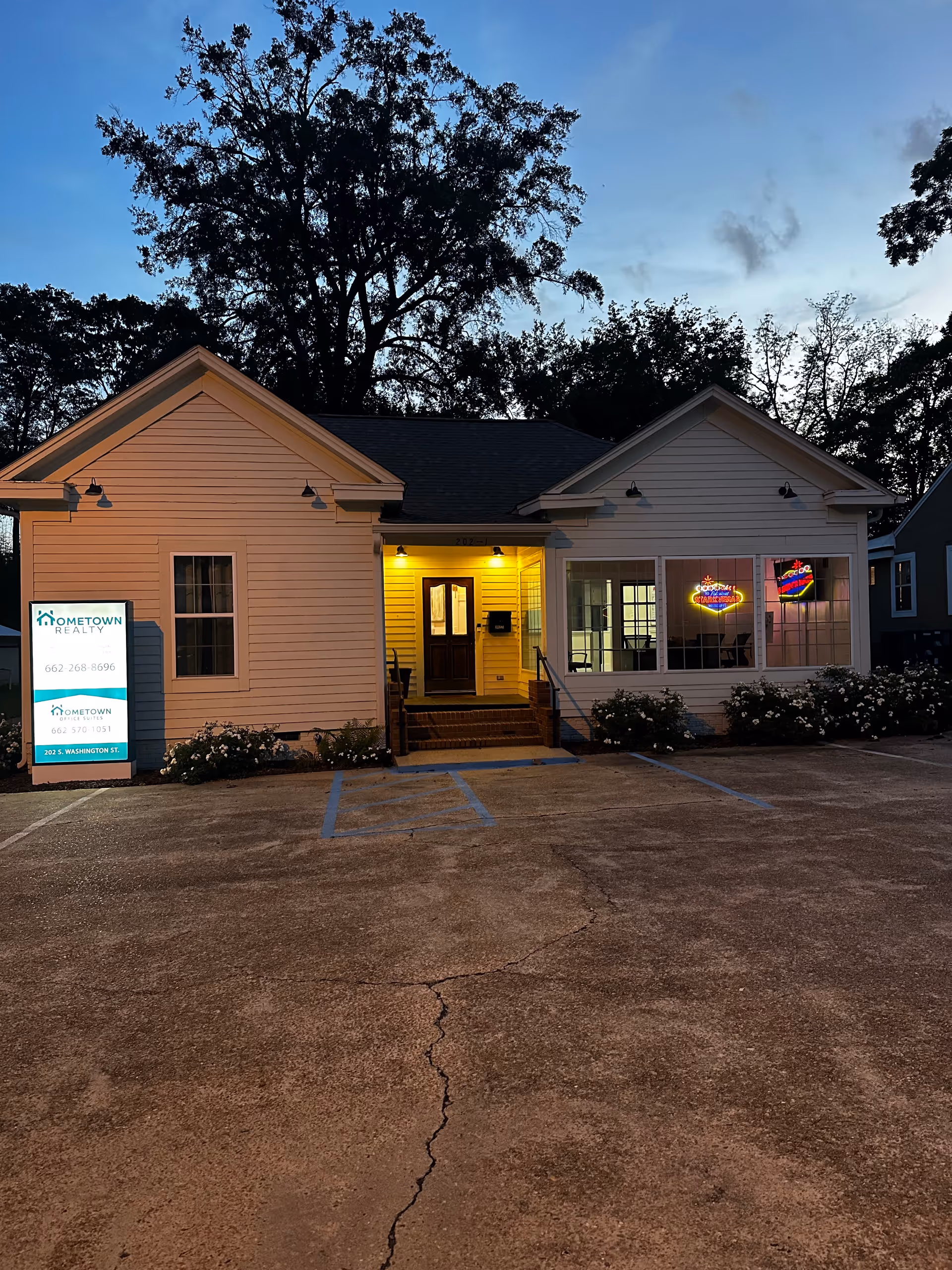 Residential-style single-story building front with illuminated entrance and parking lot at dusk.